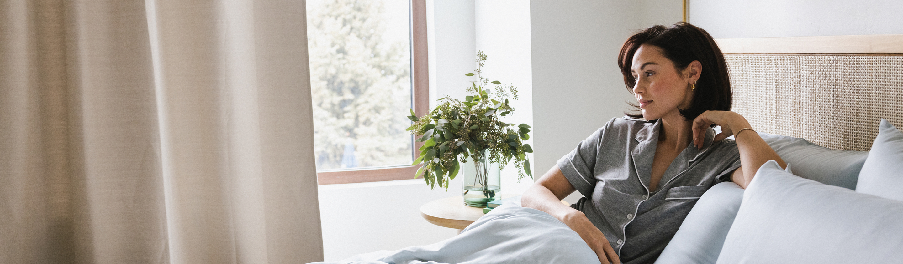 Woman in gray pajamas sits on a bed gazing out the window. Sunlight filters through beige curtains, illuminating a vase of green foliage on a wooden side table. The bedding is light blue, creating a calm and peaceful atmosphere.