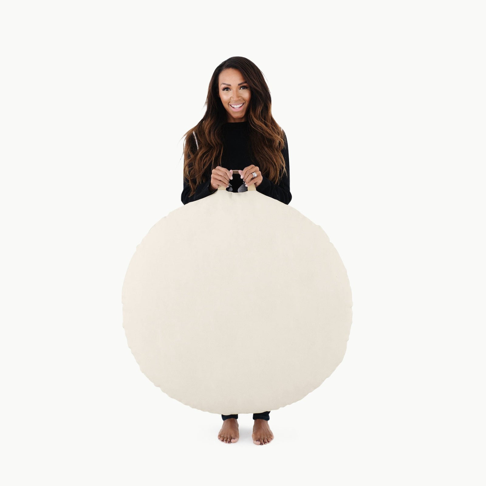 A woman with long brown hair, dressed in black and barefoot, stands against a plain white background holding the Gathre Wholesale Floor Cushion in Ivory, partially covering her body.
