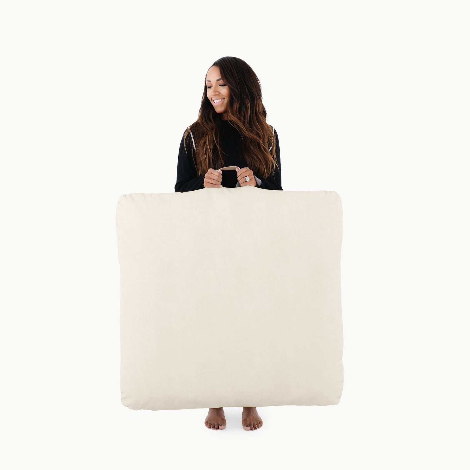 A woman with long hair stands barefoot, smiling and holding the Gathre Wholesale Floor Cushion in Ivory in front of her against a plain white background.