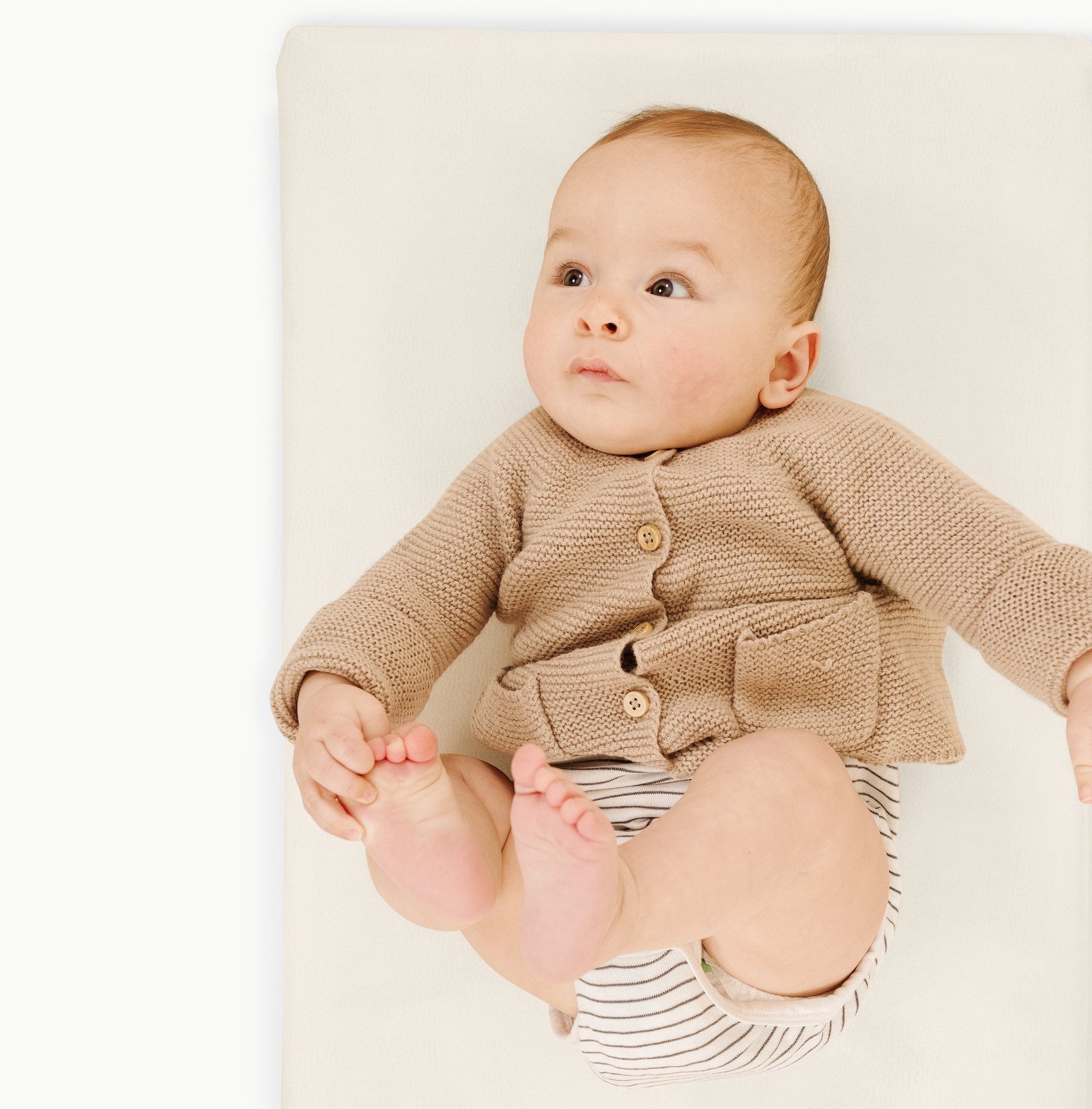 A baby lies on a cream Wholesale Padded Changing Mat by Gathre, wearing a beige knitted sweater and striped diaper cover, gazing upward with curiosity.