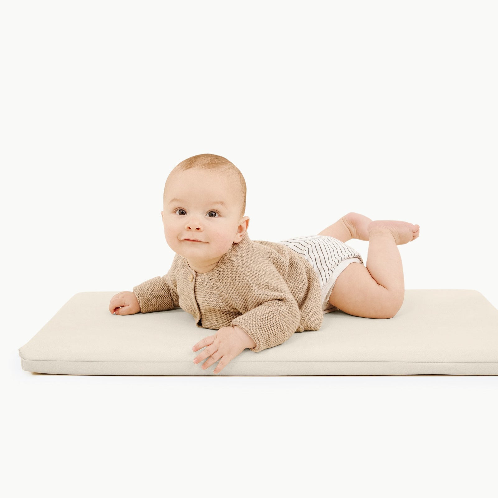 A baby lies on their stomach on an ivory Gathre Wholesale Padded Changing Mat, wearing a beige sweater and striped shorts, set against a plain white background.
