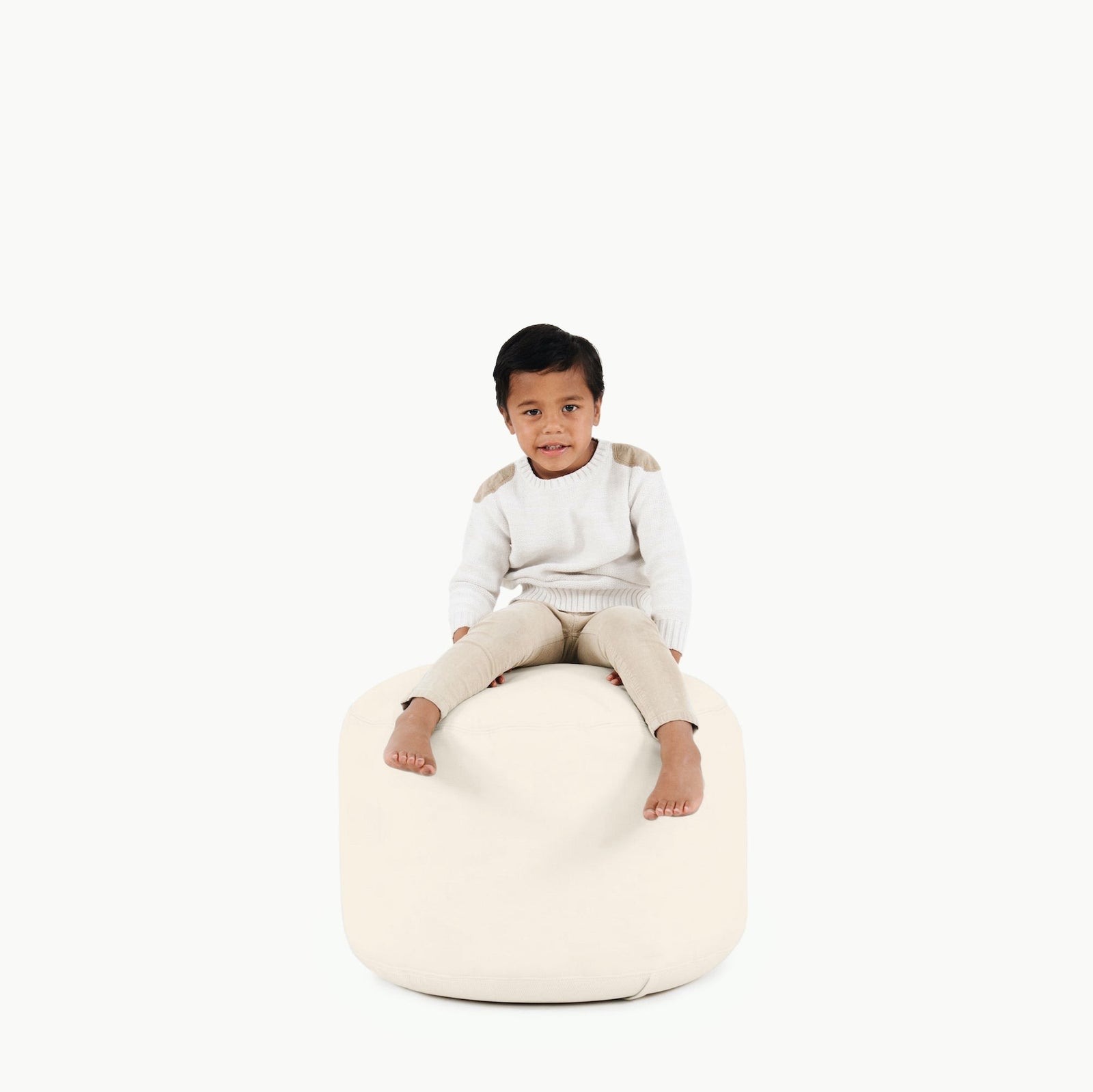 A young boy sits barefoot on the Gathre Wholesale Pouf in Ivory, placed against a plain white background. He wears a white sweater with beige accents and beige pants, looking forward with a slight smile.
