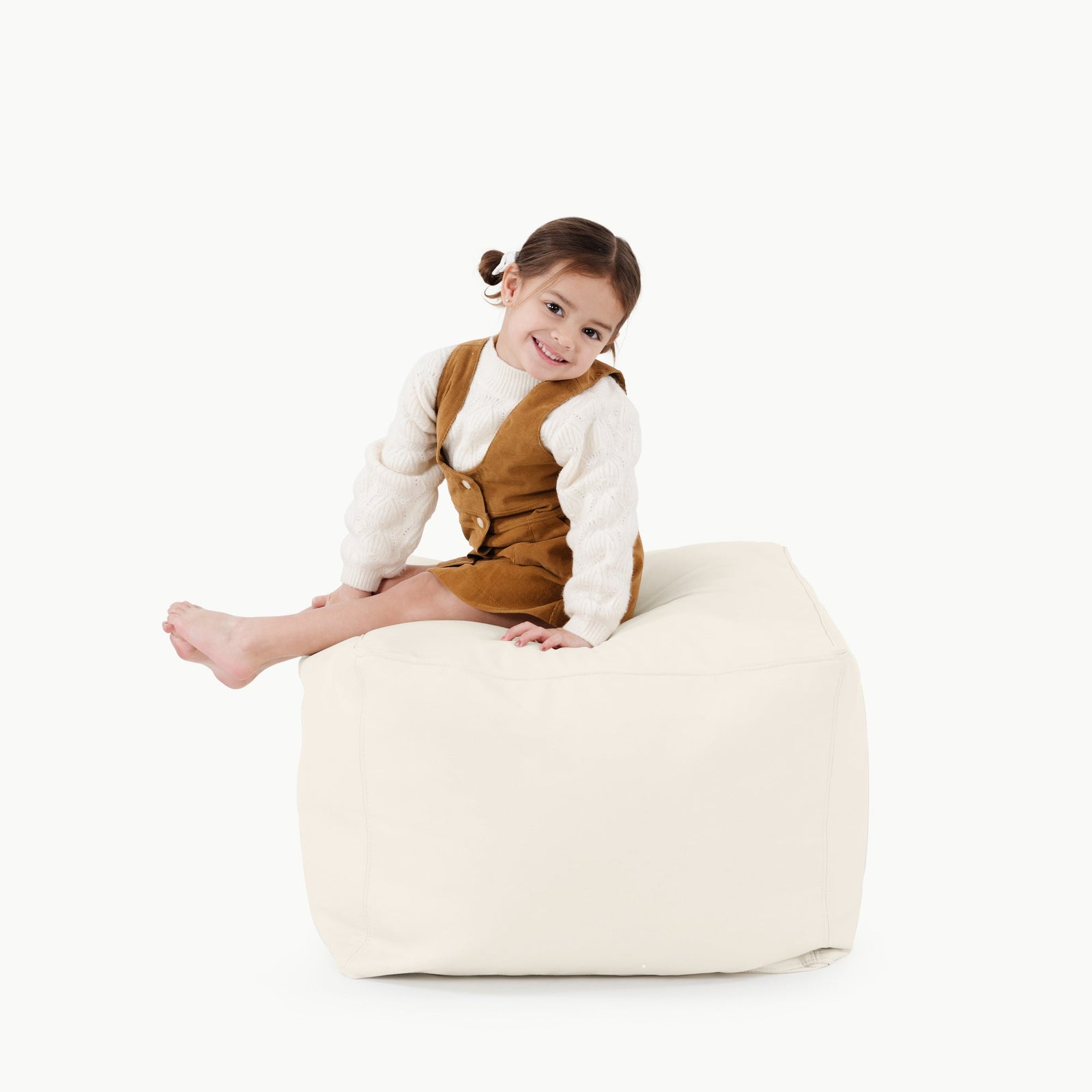 A young girl with brown hair in pigtails, wearing a white sweater and brown overalls, sits barefoot and smiling on the Gathre Wholesale Pouf in Ivory against a plain white background.