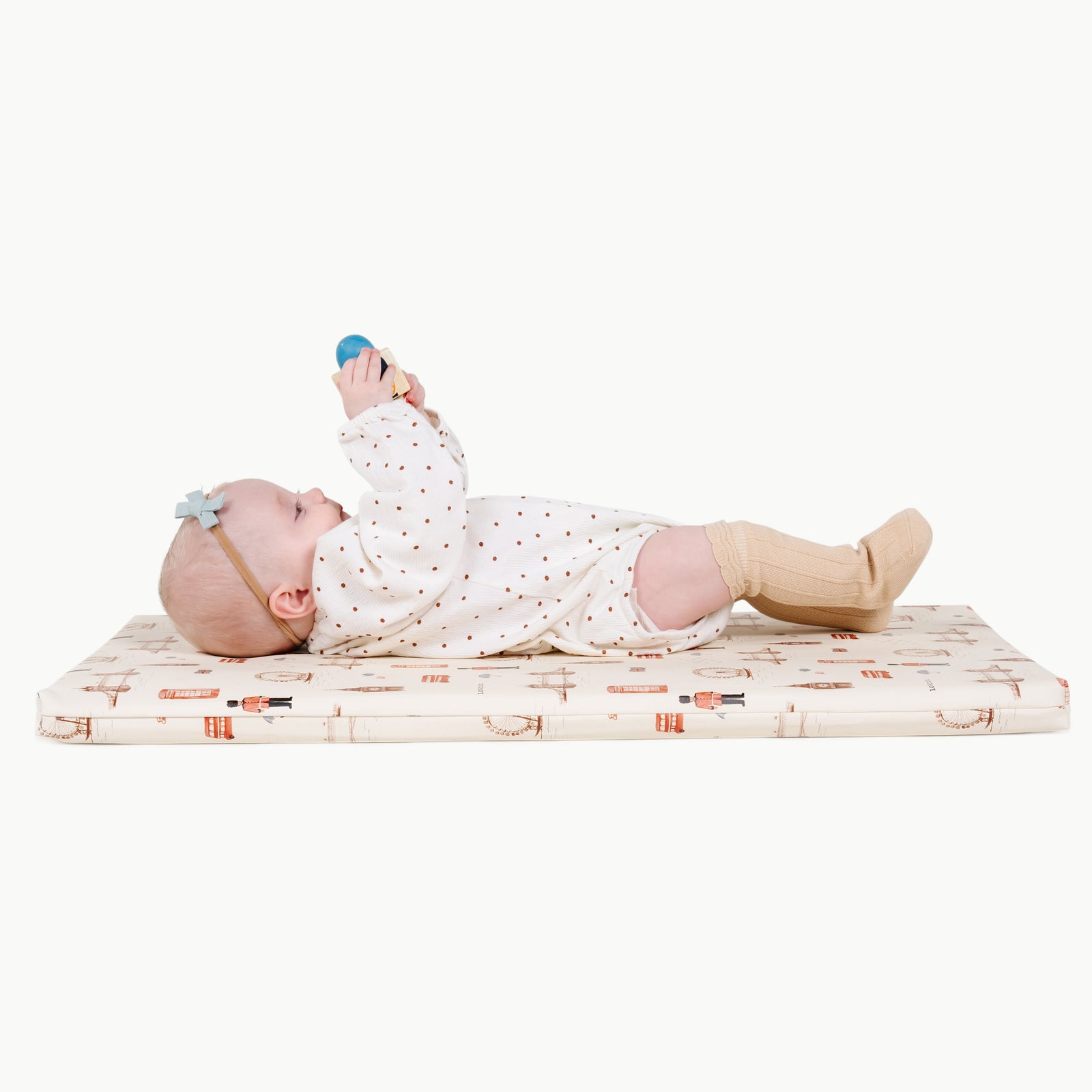 A baby in a white polka dot onesie lies on the Gathre Wholesale Padded Changing Mat in London color, holding a small blue object. The background is white.