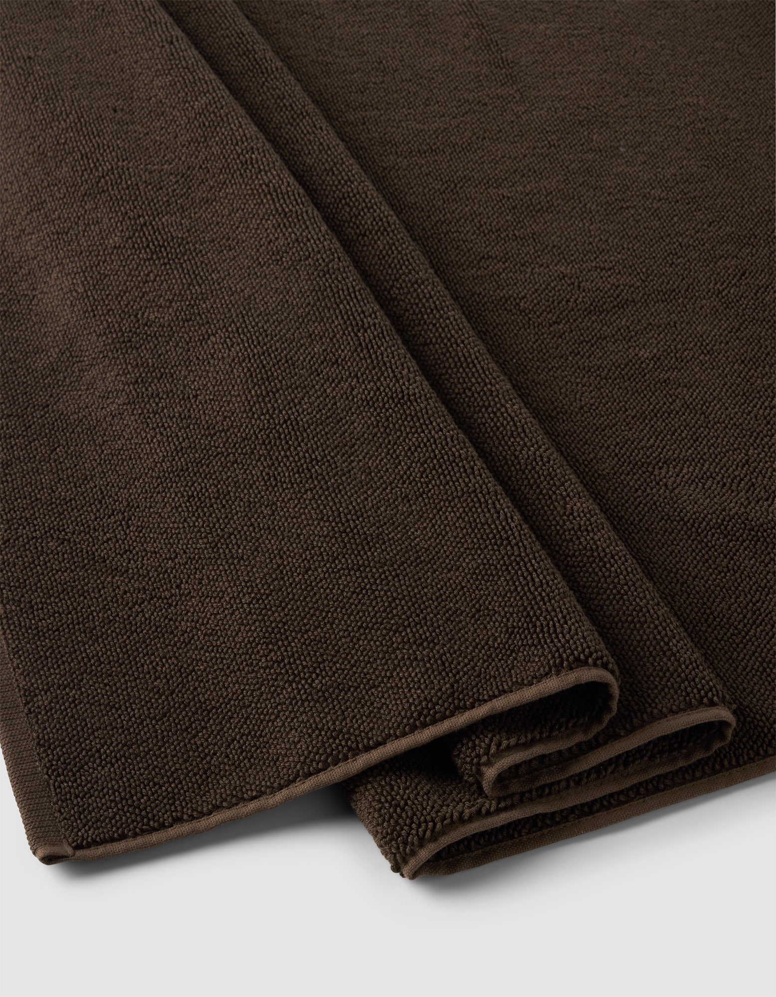A close-up of two folded Cozy Earth Loop Terry Bath Mats in dark brown, featuring textured woven fabric, placed on a light gray surface.