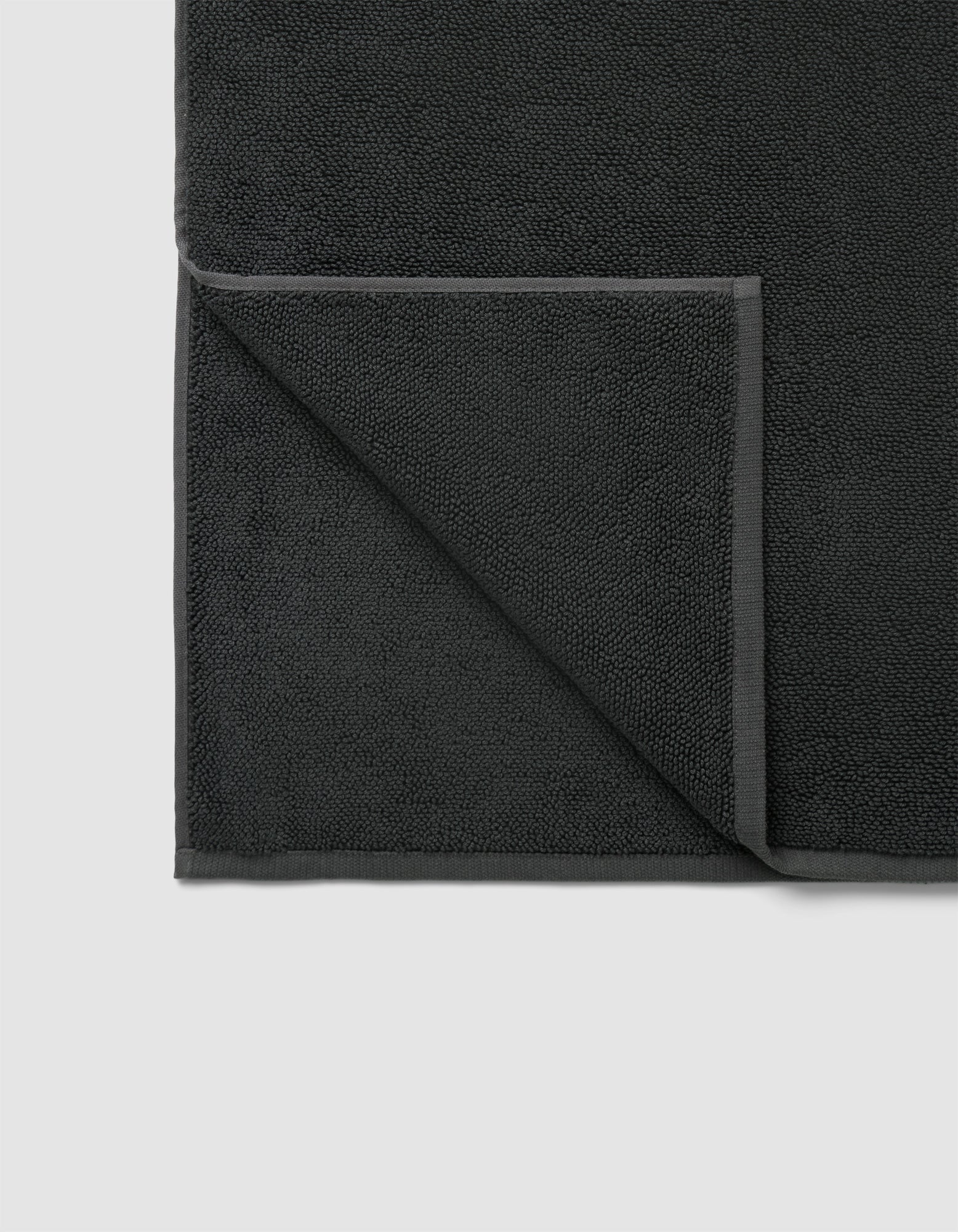 The Cozy Earth Loop Terry Bath Mat in dark gray is pictured neatly folded at one corner, displaying its textured surface and smooth edge border on both front and back sides.
