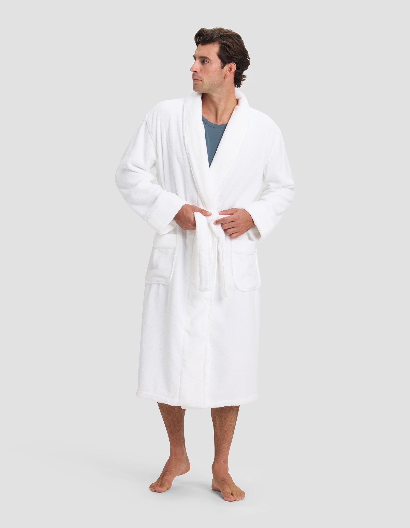 A man with short, dark hair stands barefoot in a Cozy Earth Luxe Bath Robe over a blue shirt, tying the belt at his waist. He faces slightly right against a plain light gray background.