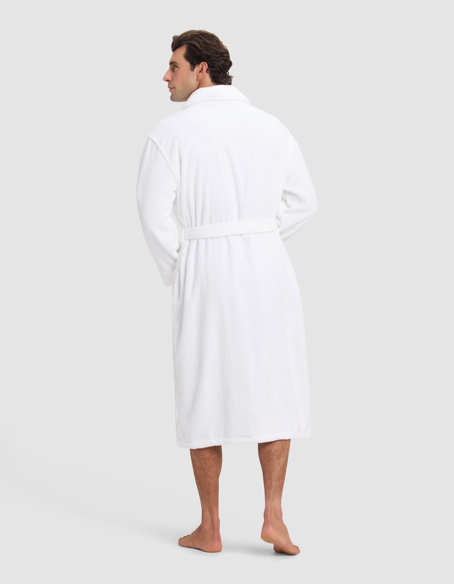 A man stands barefoot facing away, wearing the Luxe Bath Robe by Cozy Earth, featuring a belt, against a plain light gray background.
