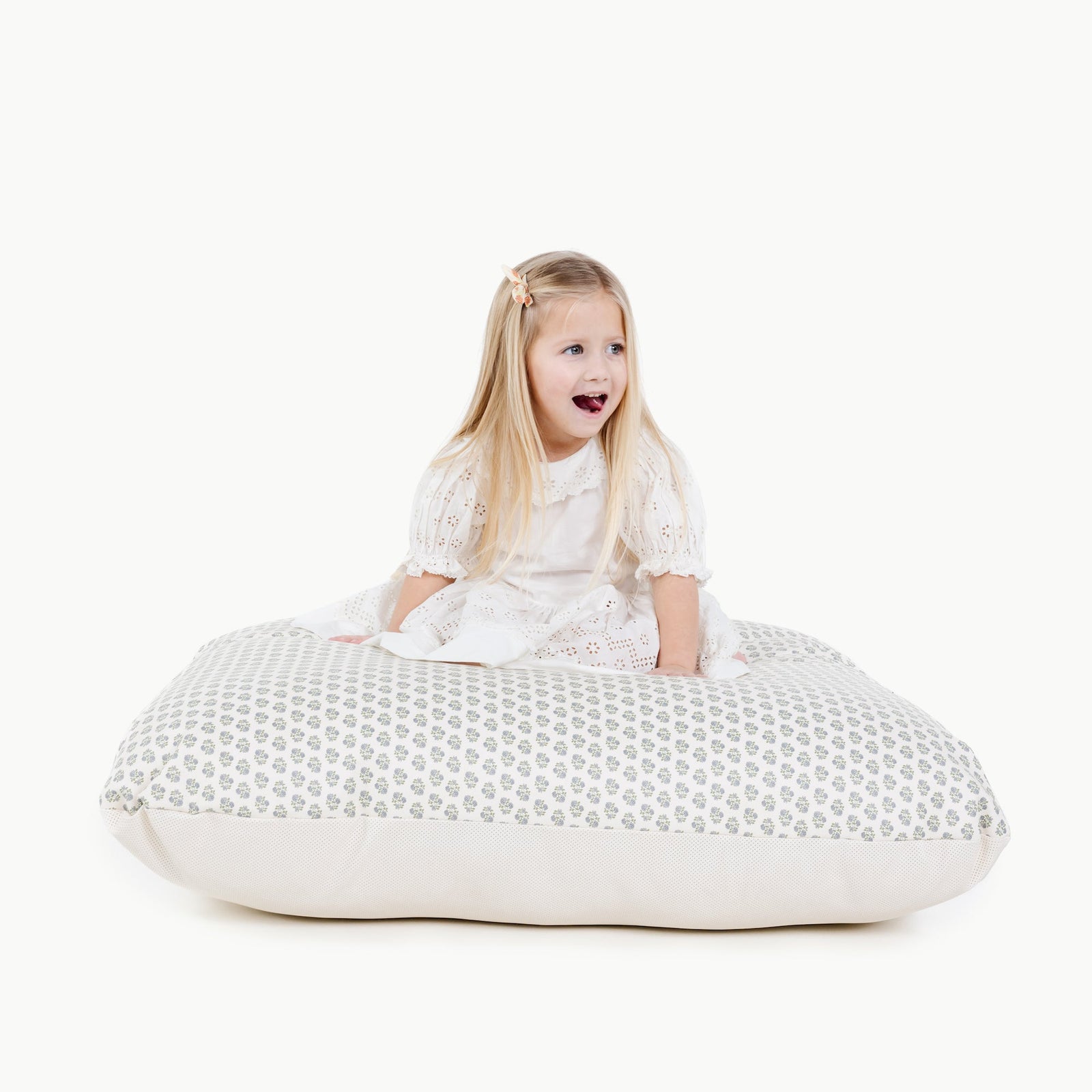 A young girl with long blonde hair, wearing a white dress, sits on a large Gathre Wholesale Floor Cushion against a plain white background, looking surprised or excited with her mouth open and eyes wide.