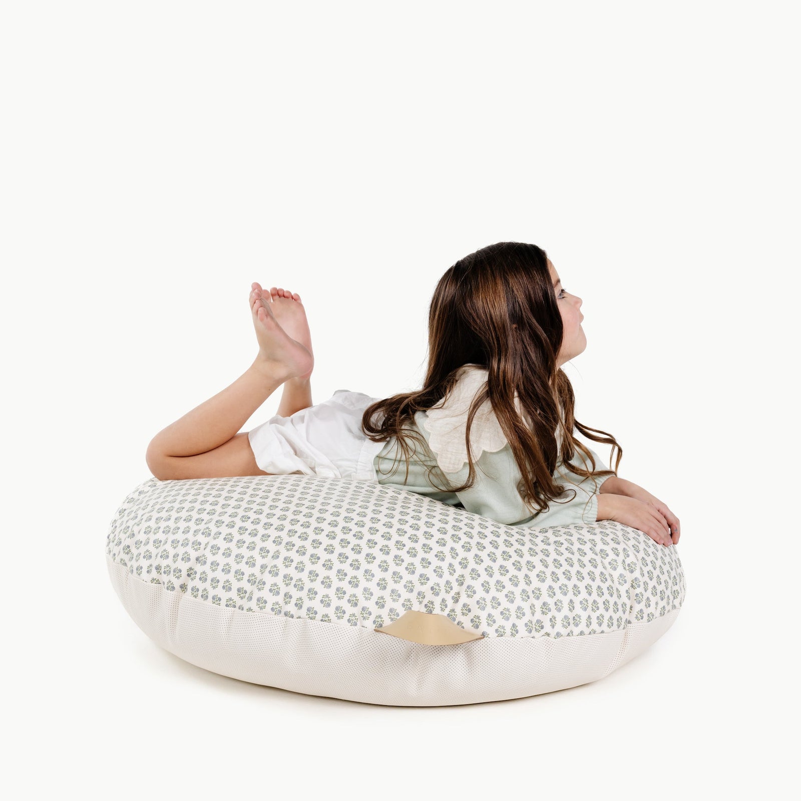 A young girl with long brown hair lies on her stomach atop a Gathre Wholesale Floor Cushion, legs bent and feet up, set against a plain white background.