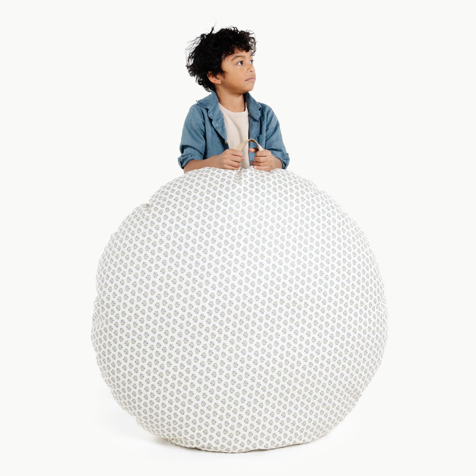 A child with curly hair and a denim shirt stands behind the Gathre Wholesale Floor Cushion in Meadow, resting their hands on top and looking to the side against a plain white background.