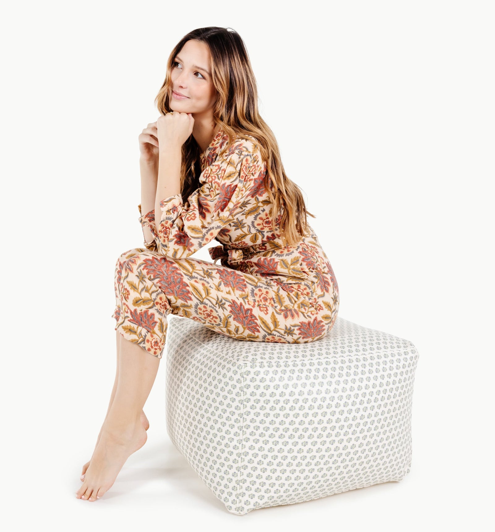 A woman in floral-patterned pajamas sits barefoot on a Gathre Wholesale Pouf in Meadow, resting her chin on her hands and gazing thoughtfully to the side against a white background.