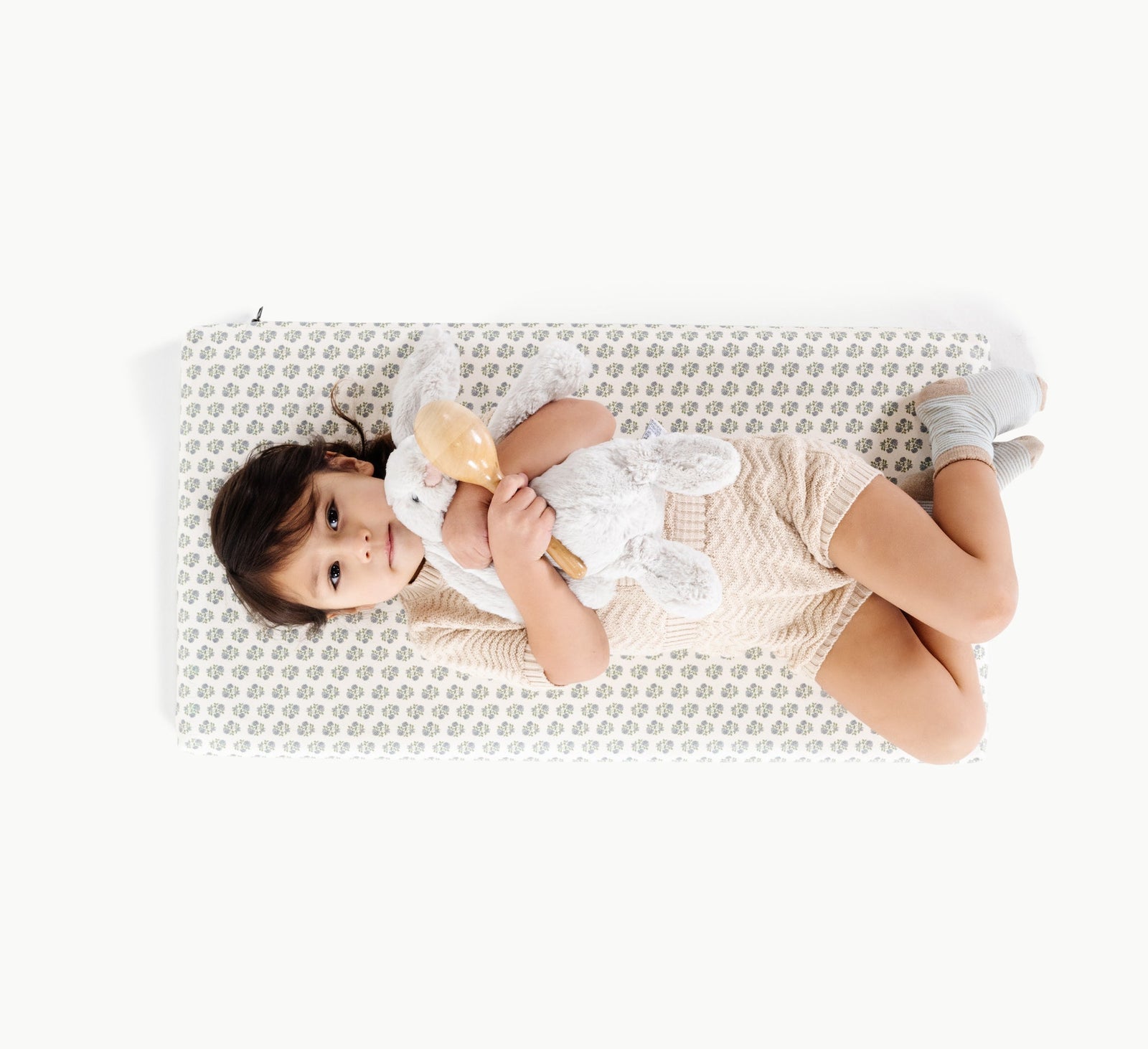 A young child lies on a Gathre Wholesale Padded Changing Mat, hugging a white stuffed bunny and a wooden rattle, dressed in a beige outfit with gray socks, against a plain white background.