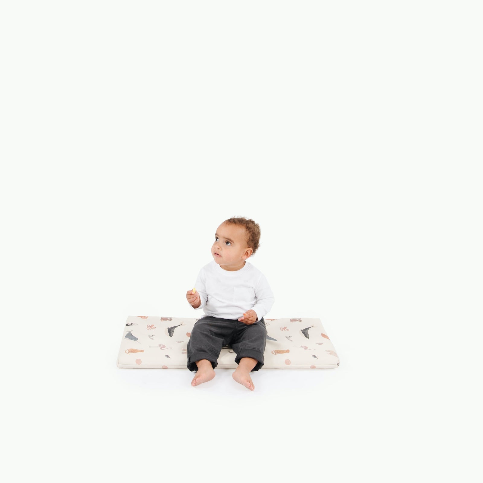A young child sits barefoot on the Gathre Wholesale Padded Changing Mat in Menagerie, against a plain white background, wearing a white long-sleeve shirt and dark pants and looking slightly upward to the right.