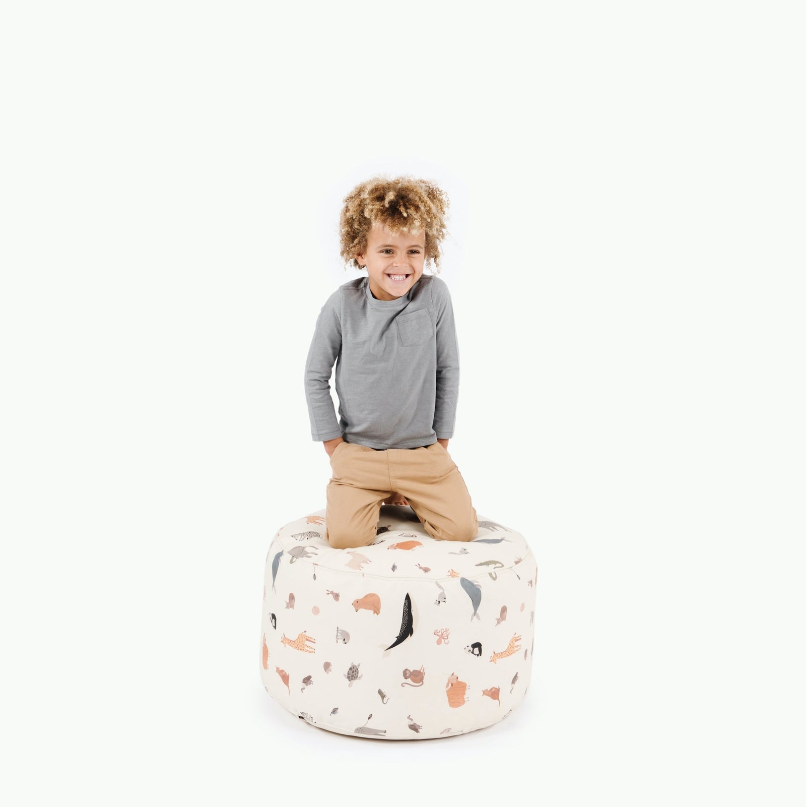 A smiling young child kneels on the Gathre Wholesale Pouf in Menagerie, featuring animal illustrations, against a plain white background.