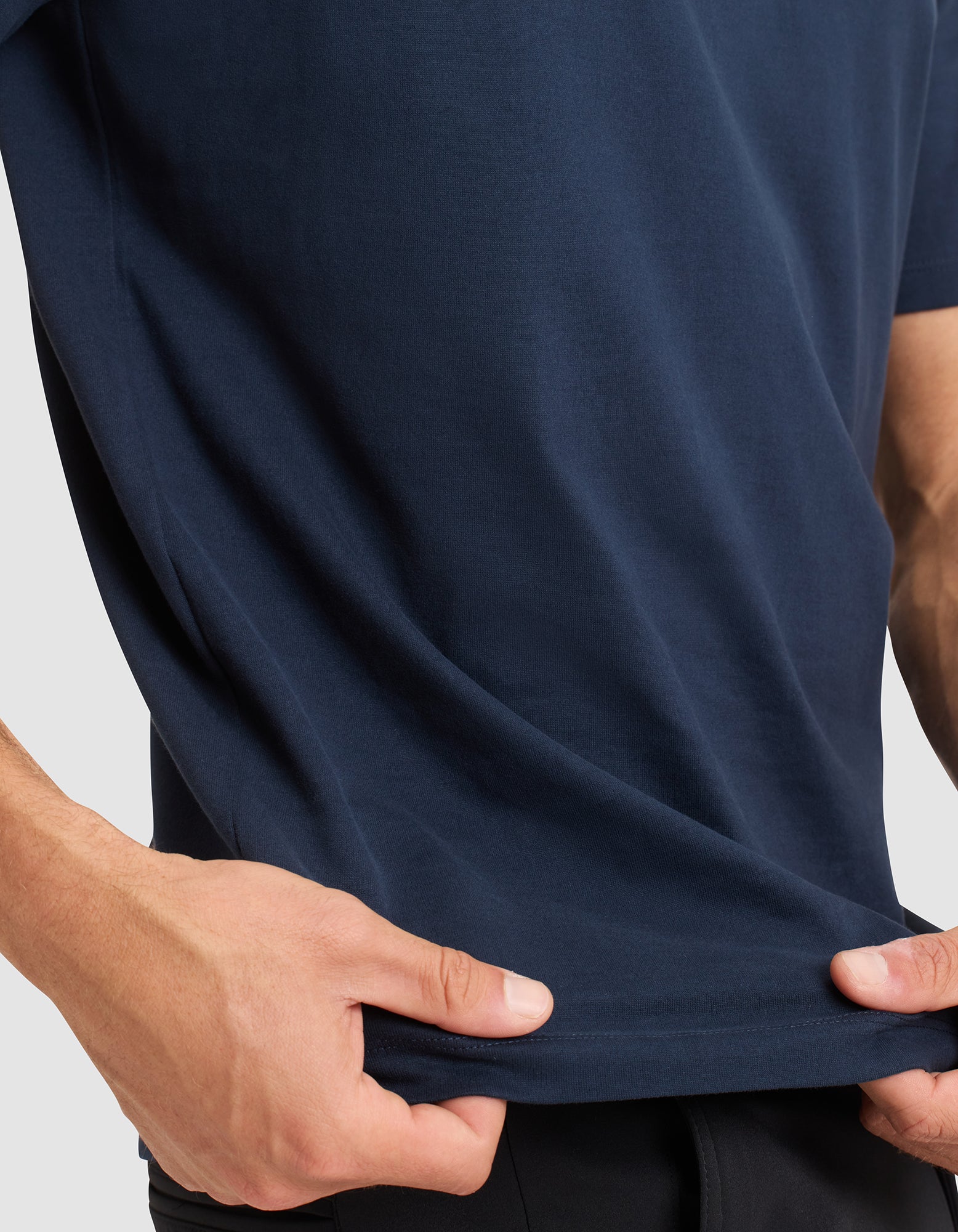 A person wearing the Cozy Earth Men's All Day Tee in navy blue holds the bottom edge of the shirt with both hands, slightly stretching the fabric; only their torso and hands are visible.