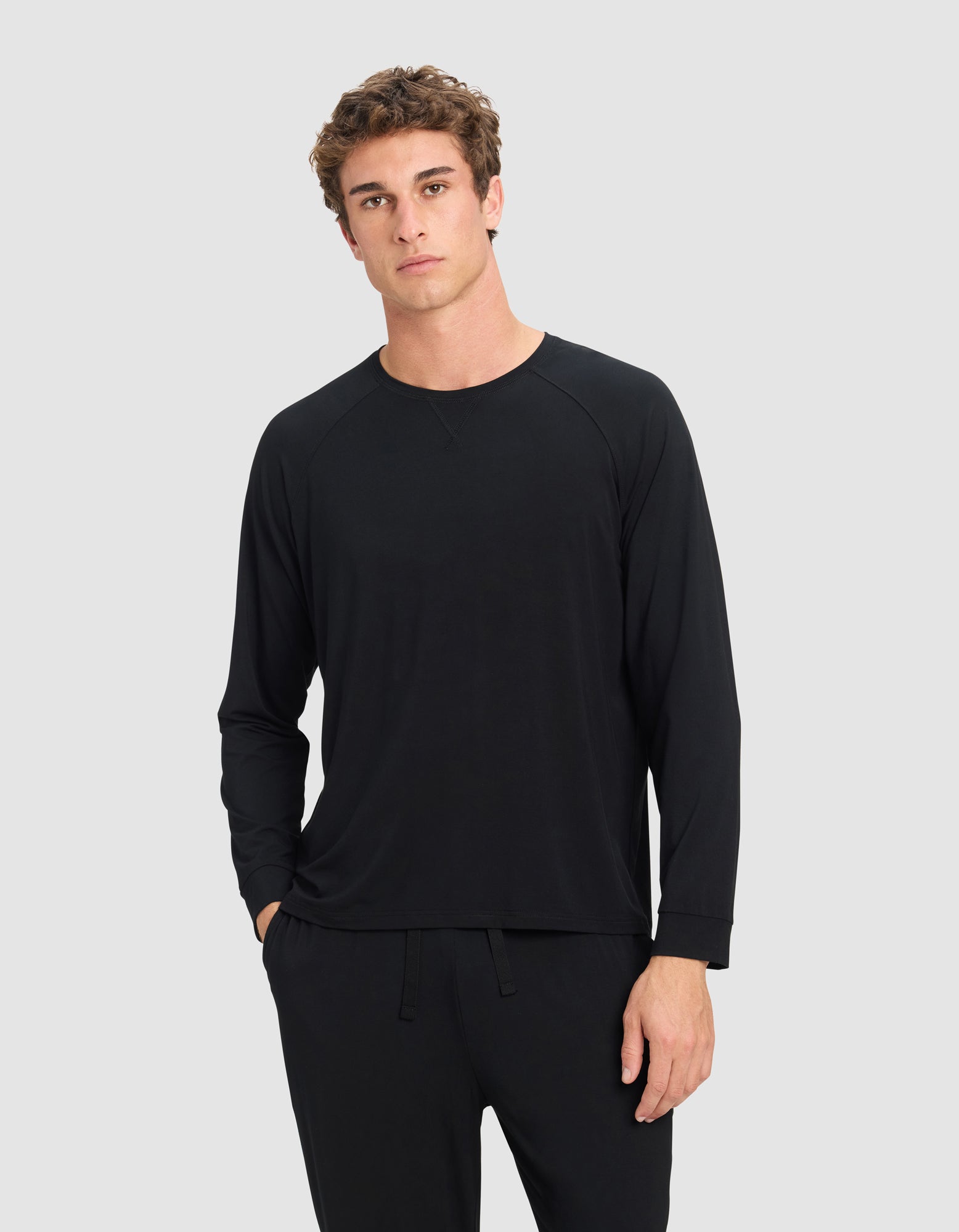 A young man with short, curly brown hair models the Cozy Earth Men's Bamboo Stretch-Knit Long Sleeve in black, paired with matching pants. He stands against a light gray background with a neutral expression and one hand in his pocket. |Color:Black