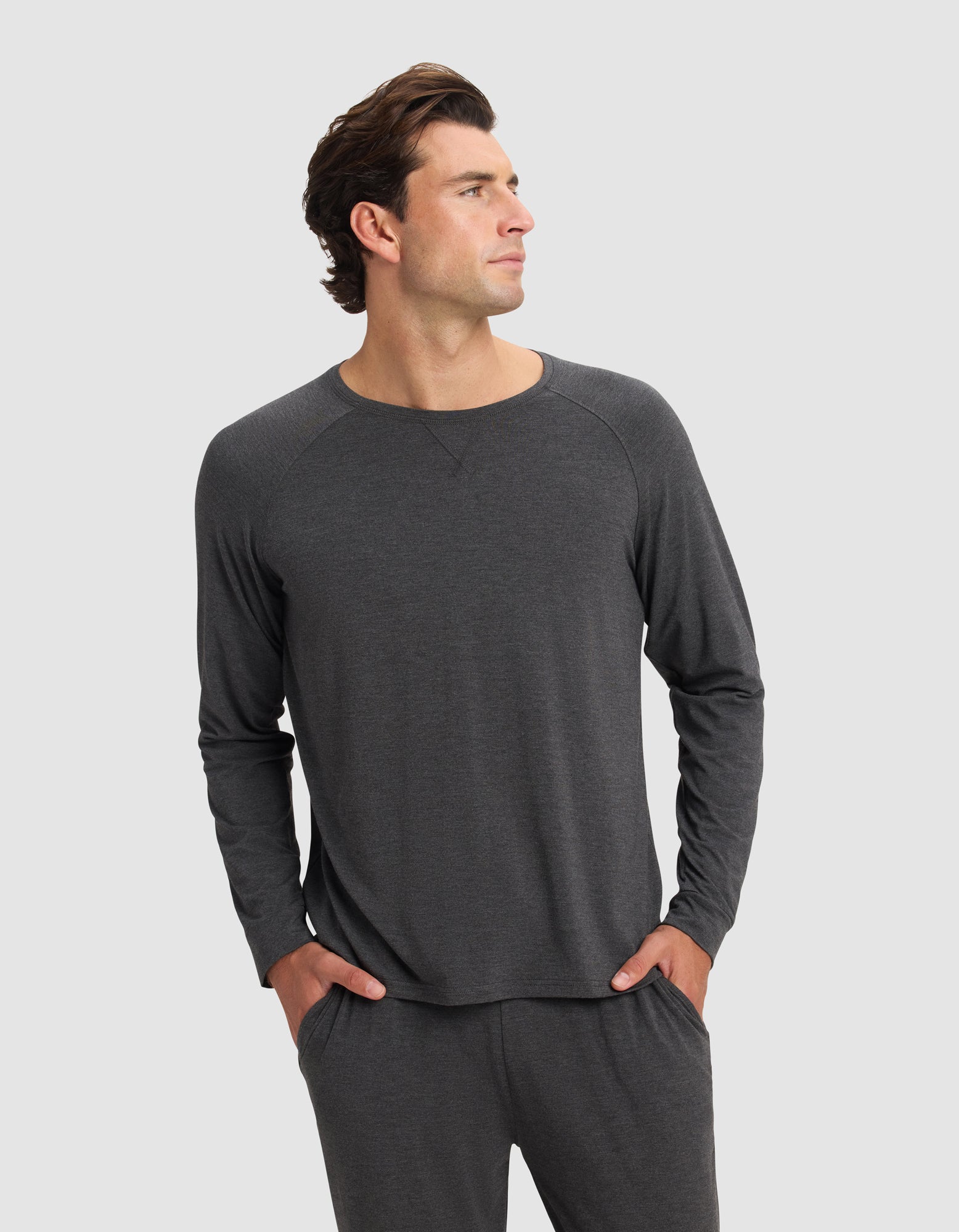 A man wearing the Cozy Earth Men's Bamboo Stretch-Knit Long Sleeve in dark gray stands with his hands in his pockets, looking to his left, against a plain light background.|Color:Charcoal