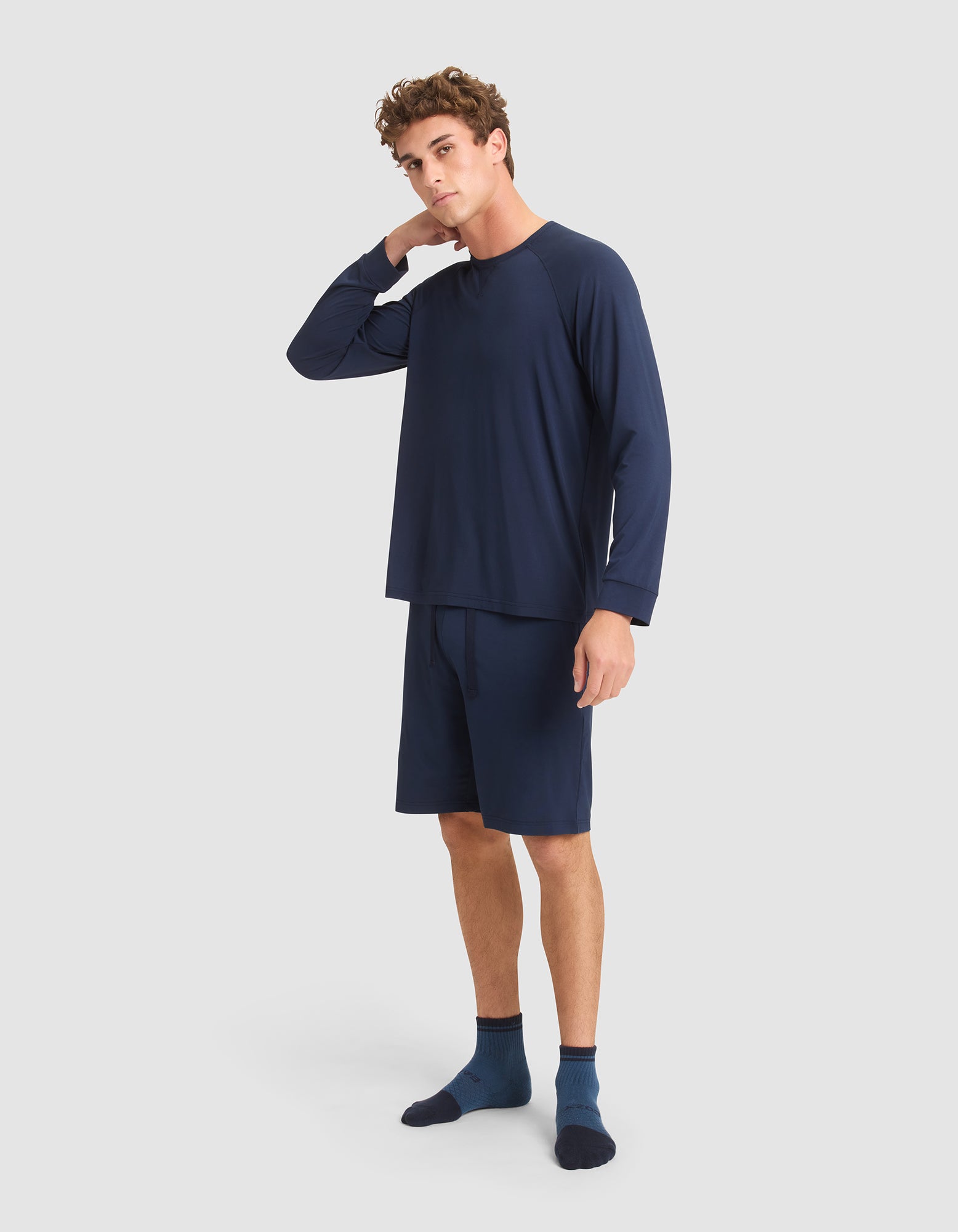 A young man stands against a plain light background, wearing the Cozy Earth Men's Bamboo Stretch-Knit Long Sleeve in navy blue. He touches the back of his neck with one hand and gazes slightly to the side. |Color:Navy