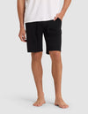 A person stands barefoot against a light gray background, wearing Cozy Earth Men's Bamboo Stretch-Knit Pajama Shorts in black with a white T-shirt. Their hands are relaxed—one in a pocket, the other by their side.|Color:Black