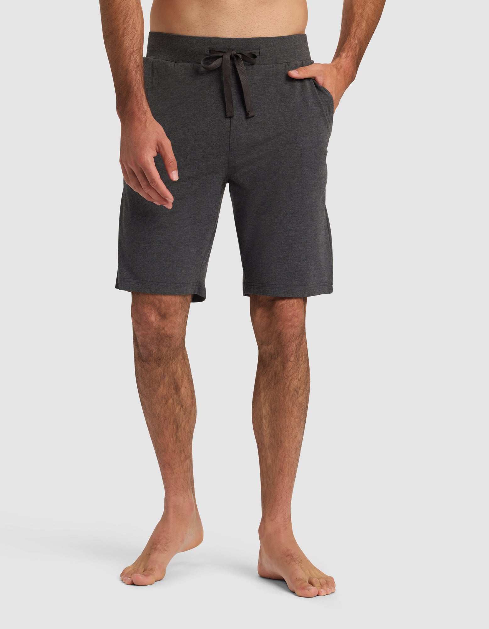A man models Cozy Earth Men's Bamboo Stretch-Knit Pajama Short in dark gray, barefoot against a plain white background, with one hand by his side and the other in his pocket.|Color:Charcoal