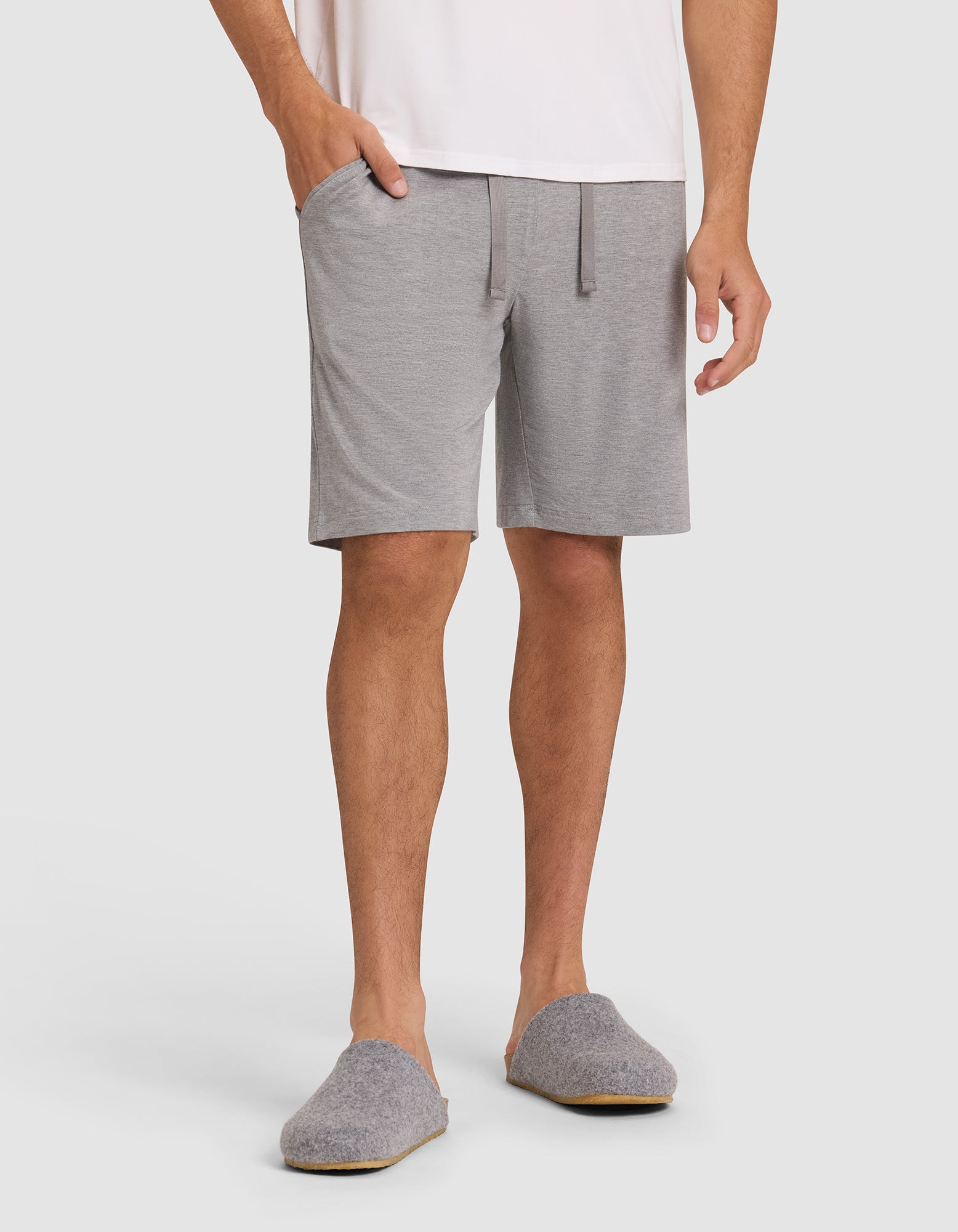 Only the lower half of a person is visible as they wear Cozy Earth's Men's Bamboo Stretch-Knit Pajama Short in light gray with pockets, paired with gray slippers and a white shirt, standing against a plain background. |Color:Heather Grey