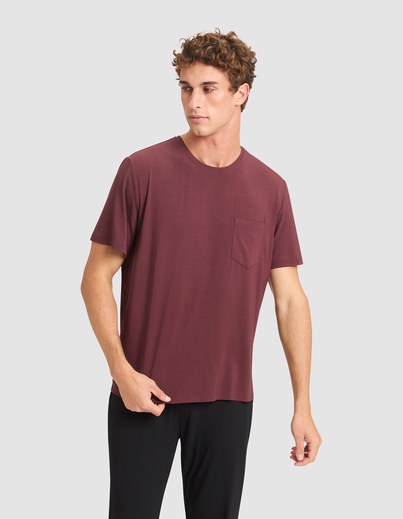 A young man in Cozy Earth's Men's Bamboo Stretch-Knit Lounge Tee and black pants stands against a gray background, looking slightly to his right with a neutral expression. |Color:Burgundy