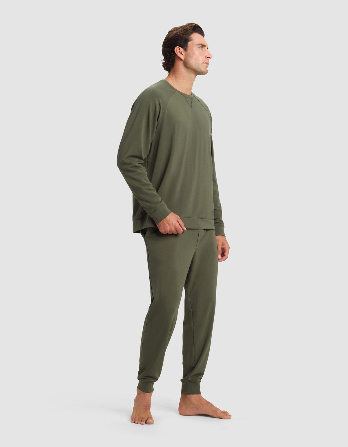 A man stands barefoot against a plain light background, wearing the Cozy Earth Men's Brushed Bamboo Jogger Set in olive green, featuring a long-sleeve top and matching pants. He faces sideways with a relaxed posture.