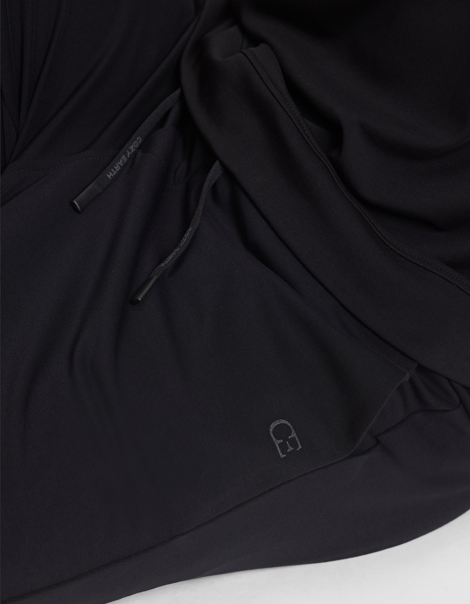 A close-up of black fabric on the Cozy Earth Men's Brushed Bamboo Jogger, featuring drawstrings—one end reads "Kindly"—and a small, subtle embroidered logo near the bottom.