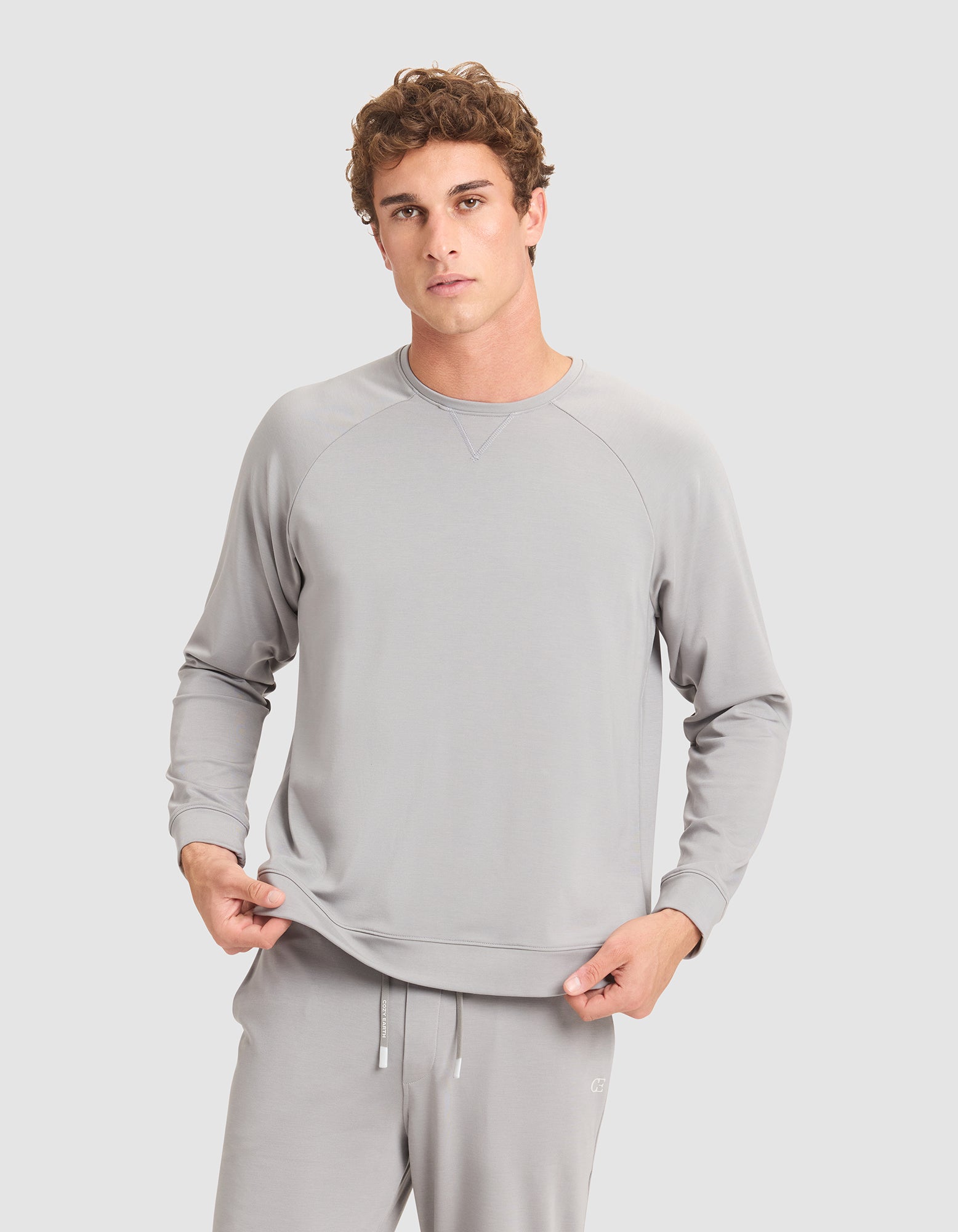A young man with curly hair wears Cozy Earth's Men's Brushed Bamboo Pullover Crew in light gray, standing against a plain gray background and holding the hem of his sweatshirt. |Color:Stone