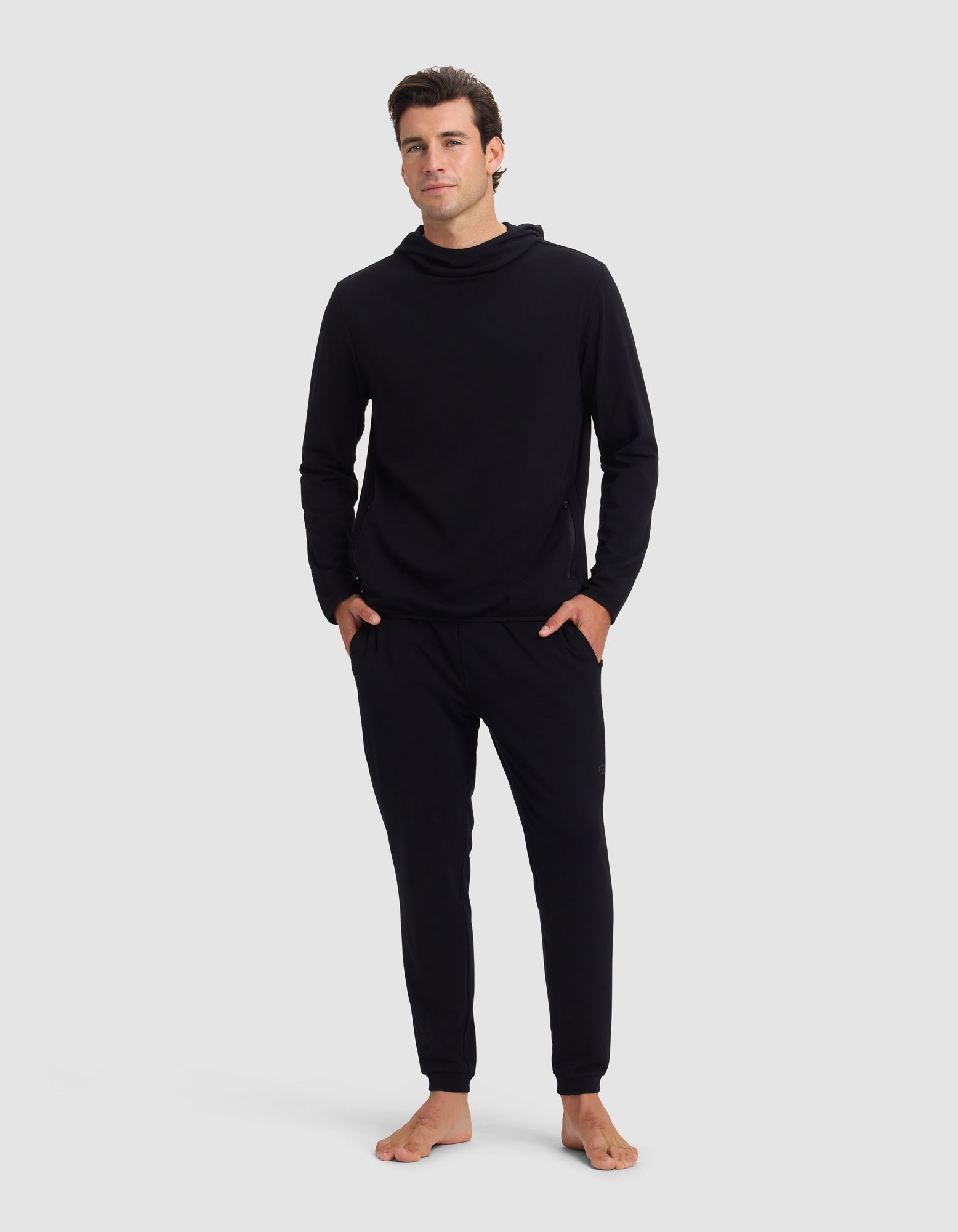 A man stands barefoot against a plain background, wearing the Cozy Earth Men's Brushed Bamboo Recovery Hoodie & Jogger Set in black. His hands are in his pockets and he gazes forward with a neutral expression.|Color:Black