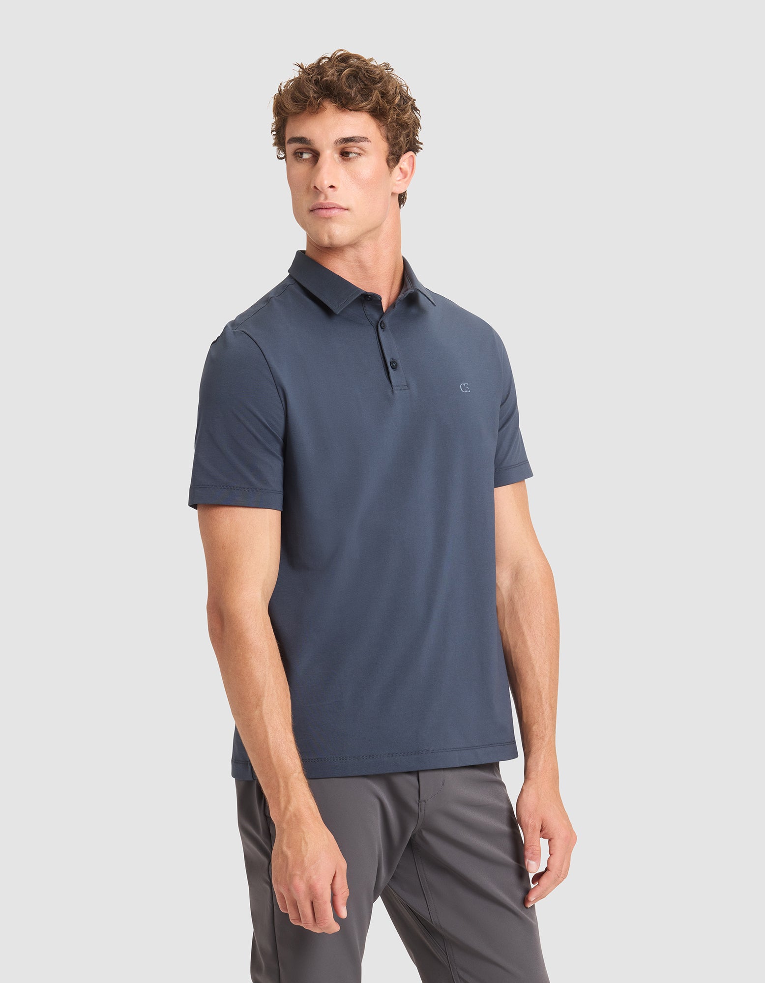 A man with short curly hair wears the Cozy Earth Men's Everyday Polo in dark blue and gray pants, standing against a plain light gray background and looking slightly left with a neutral expression. |Color:Eclipse