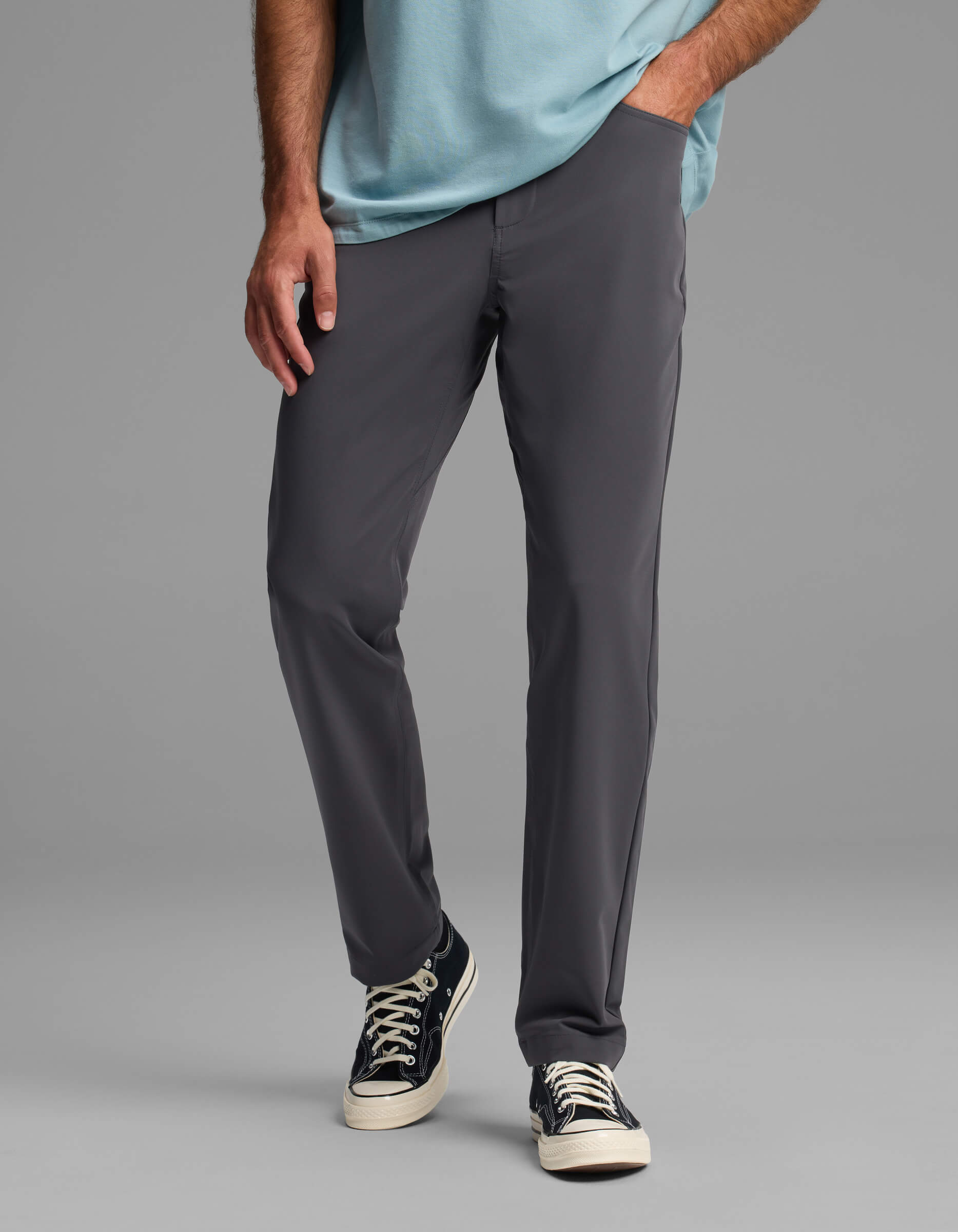 A man wears Cozy Earth's Men's Everywhere Pant - Relaxed Fit in gray, paired with a light blue shirt and black canvas sneakers with white laces, standing with one hand in his pocket against a plain gray background.|Color:Graphite