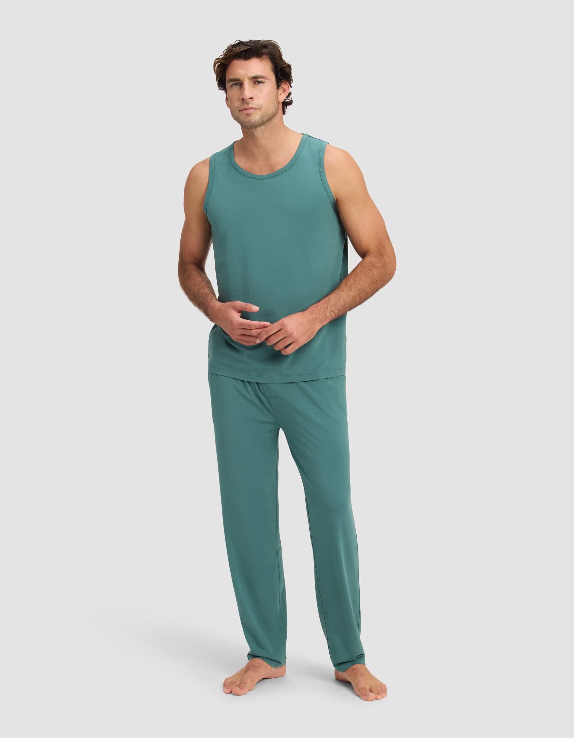 A man stands barefoot against a plain background, wearing the Cozy Earth Men's First Layer Pajama Tank & Pant Set in teal. He faces forward with a relaxed expression, one hand touching the other.|Color:Sea Green