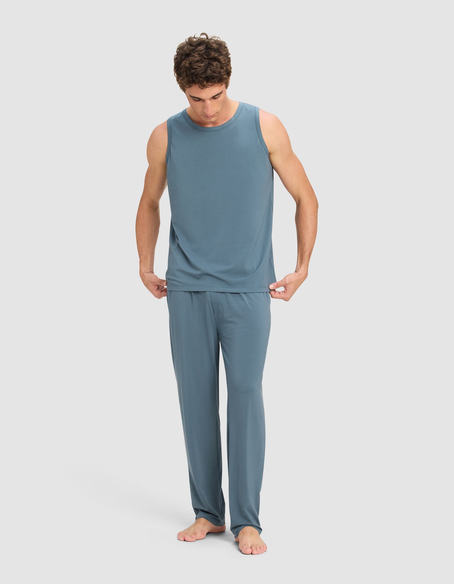 A man stands barefoot on a light background, wearing the Cozy Earth Men's First Layer Pajama Tank & Pant Set in blue. He looks down while holding the hem of his shirt with both hands.