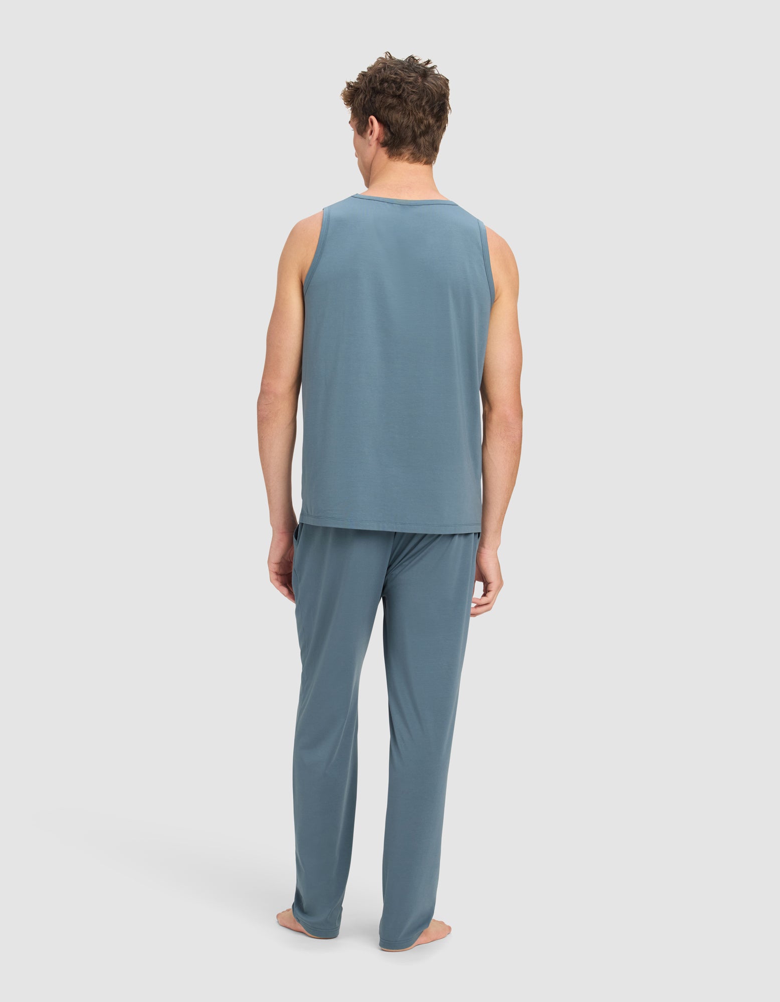 A man stands barefoot facing away, wearing the Cozy Earth Men's First Layer Pajama Tank & Pant Set in blue against a plain light background.