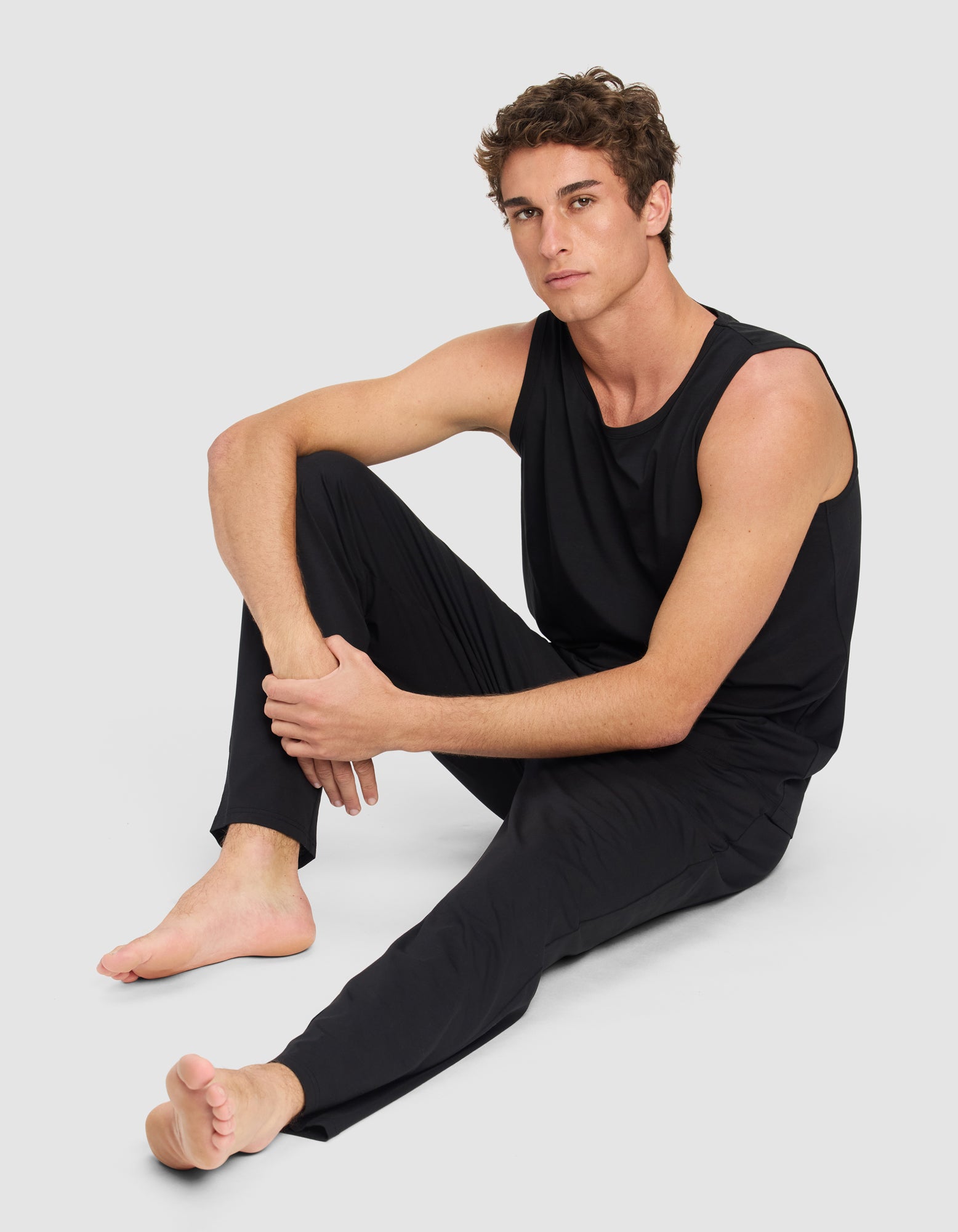 A man with short curly hair sits on the floor against a light gray background, wearing the Cozy Earth Men's First Layer Pajama Tank & Pant Set in black. He is barefoot, with one arm on his knee, looking at the camera.