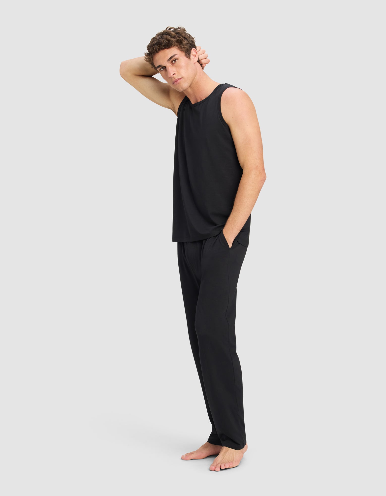 A man models the Cozy Earth Men's First Layer Pajama Tank & Pant Set in black, standing barefoot on a light background with one hand on his head and the other in his pocket.