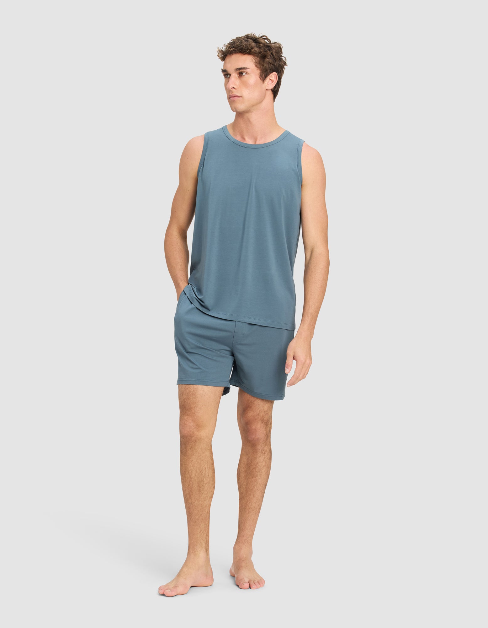 A man models the Men's First Layer Pajama Tank & Short Set by Cozy Earth, standing barefoot against a plain light gray background with one hand in his pocket, looking to the side.