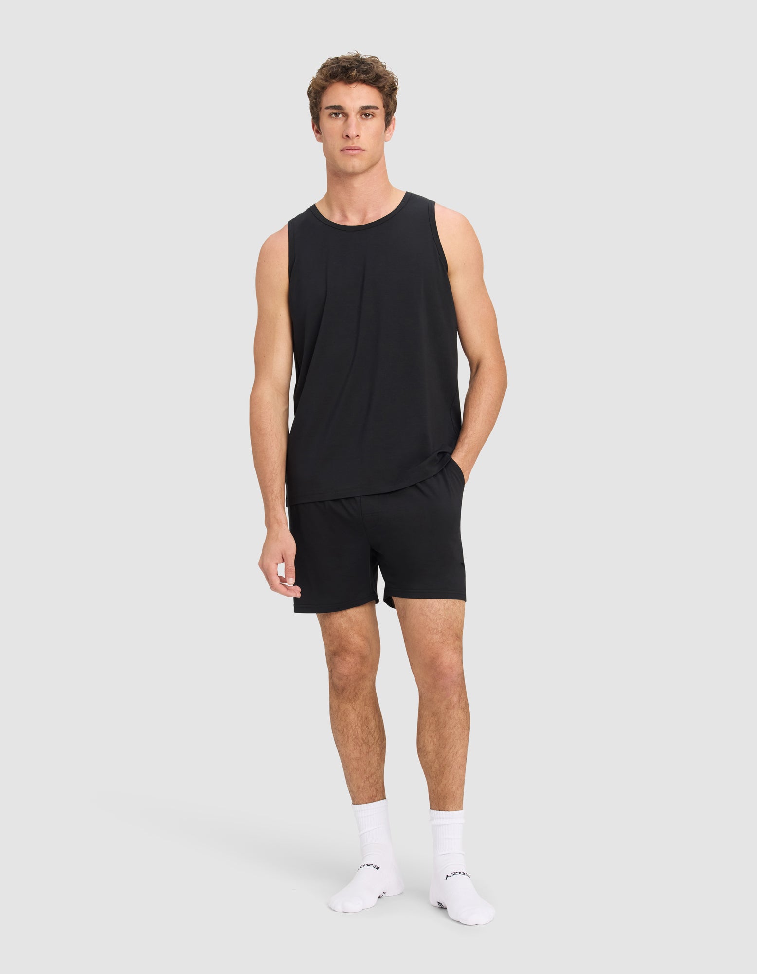 A man stands against a plain light gray background, wearing the Cozy Earth Men's First Layer Pajama Tank & Short Set in black, paired with white socks. One hand is in his pocket and he looks straight ahead with a neutral expression.