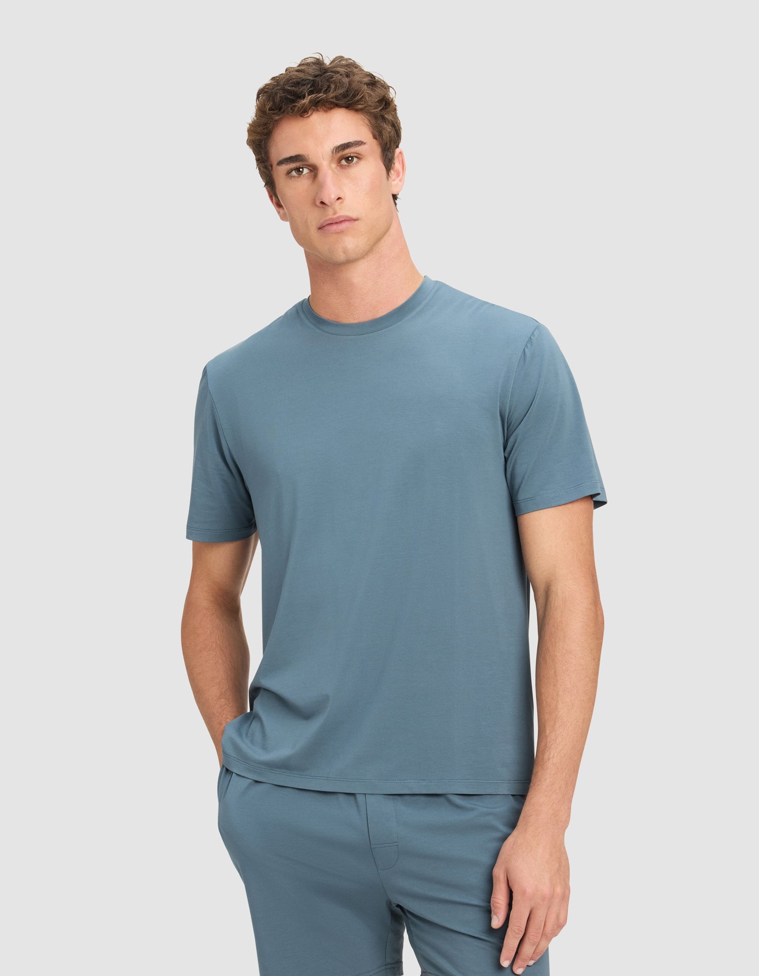 A young man with short curly hair wears the Cozy Earth Men's First Layer Pajama Short Sleeve Tee and matching shorts, standing in front of a plain light gray background with one hand in his pocket.|Color:Dawn