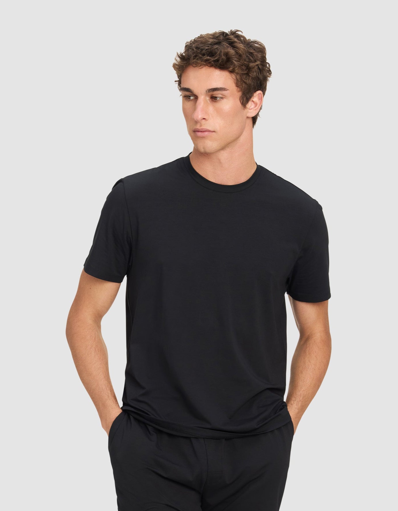 A man wears the Cozy Earth Men's First Layer Pajama Short Sleeve Tee in black, paired with black shorts, standing with hands in his pockets and looking to the side against a light gray background.|Color:Jet Black