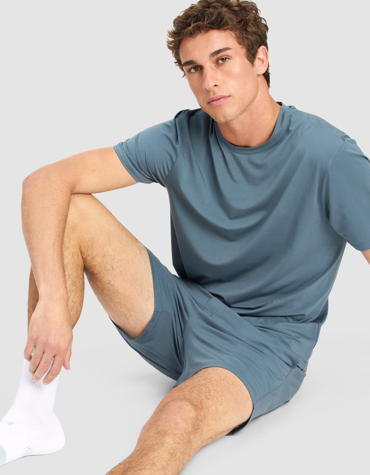 A young man with short curly hair sits on the floor wearing Cozy Earth's Men's First Layer Pajama Short Sleeve Tee & Short Set in light blue, along with white socks. One knee is up as he rests his arm on it, looking directly at the camera.