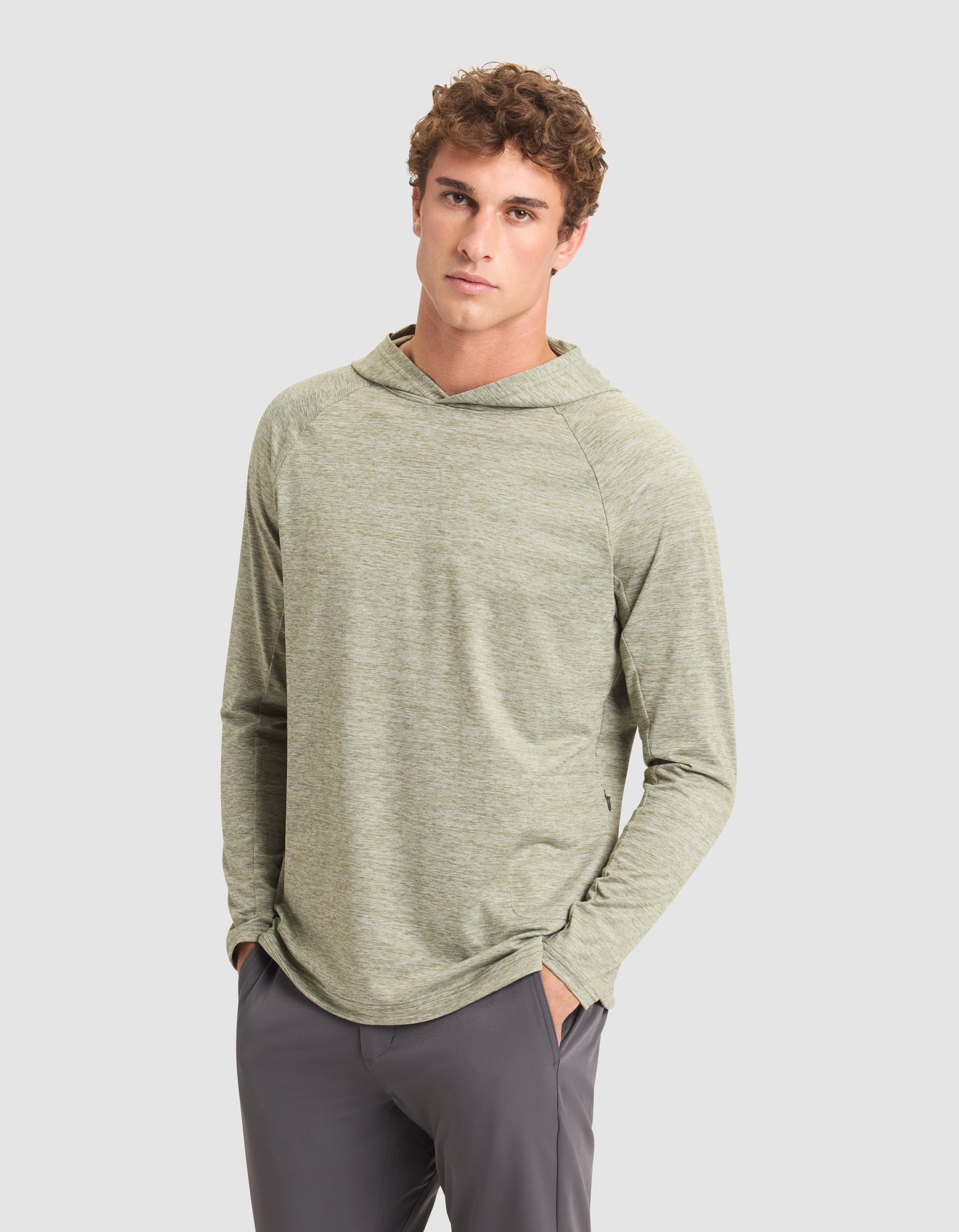 A young man with curly hair models the Cozy Earth Men's Lightweight Performance Hoodie in light green, paired with gray pants, standing against a plain light gray background with his hands in his pockets. |Color:Heather Olive