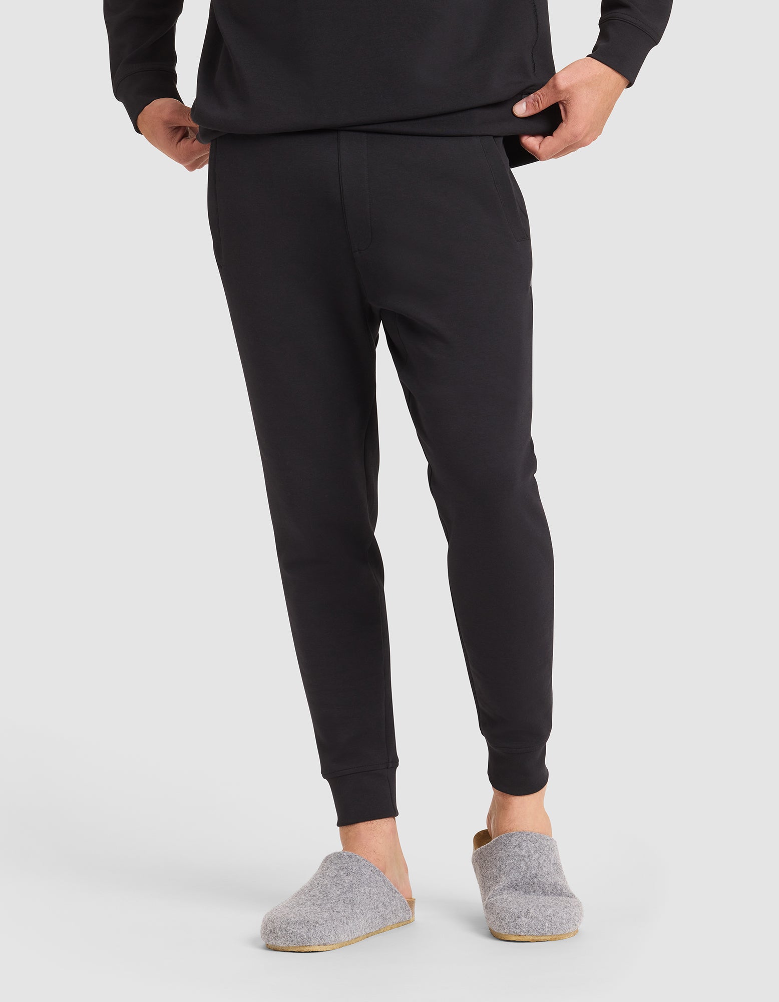 A person stands on a white background, hands in pockets, wearing Cozy Earth Men's StretchTech Jogger and a black long-sleeve top, paired with gray slip-on house slippers. |Color:Jet Black