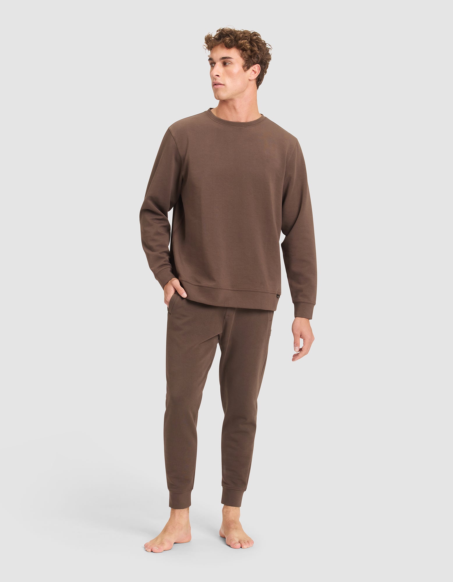 A young man with curly hair stands barefoot against a light gray backdrop, wearing the Cozy Earth Men's StretchTech Crewneck & Jogger Set in brown. |Color:Cacao