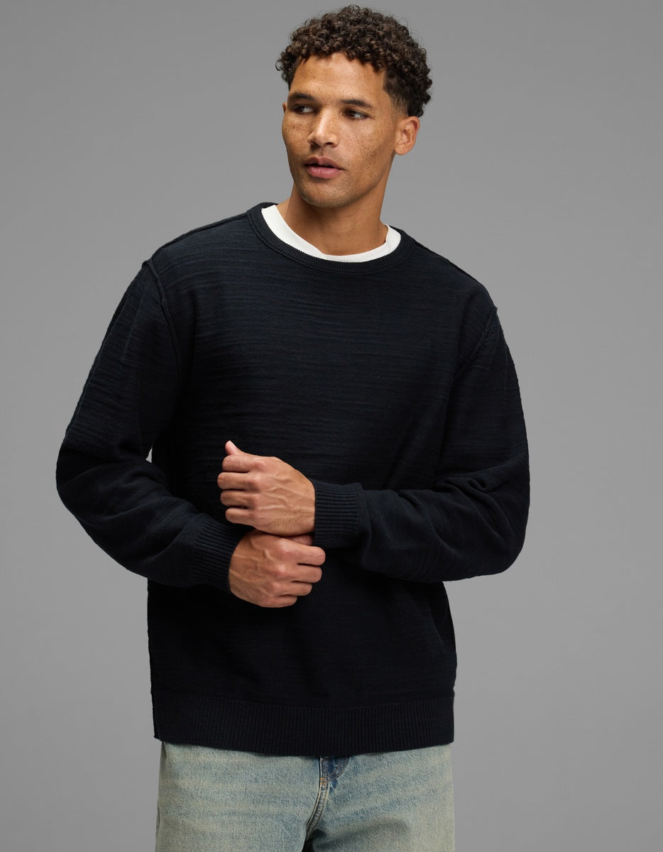 A man wears a jet black Men’s Tate Crewneck sweater with a subtle ribbed texture over a white t-shirt and light-wash jeans.|Color:Jet Black