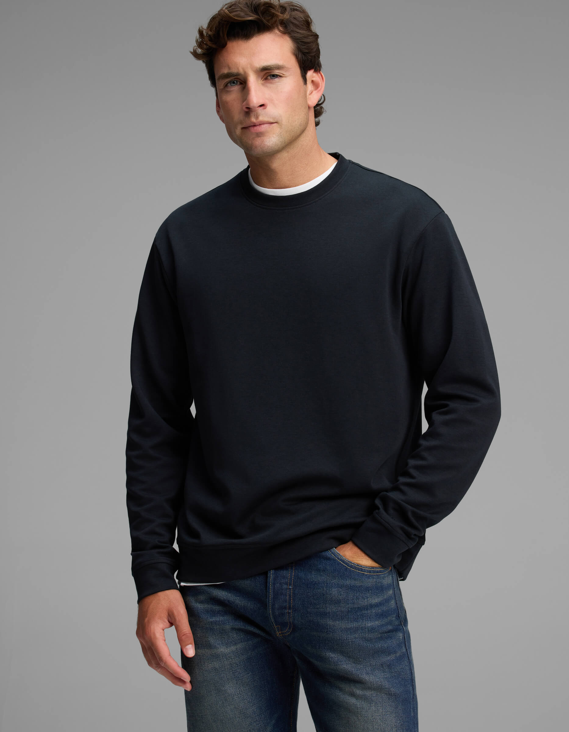 A man stands against a gray background, wearing the Cozy Earth Men's Versa Crewneck in black over a white shirt and blue jeans, with one hand in his pocket and a neutral expression.|Color:Jet Black