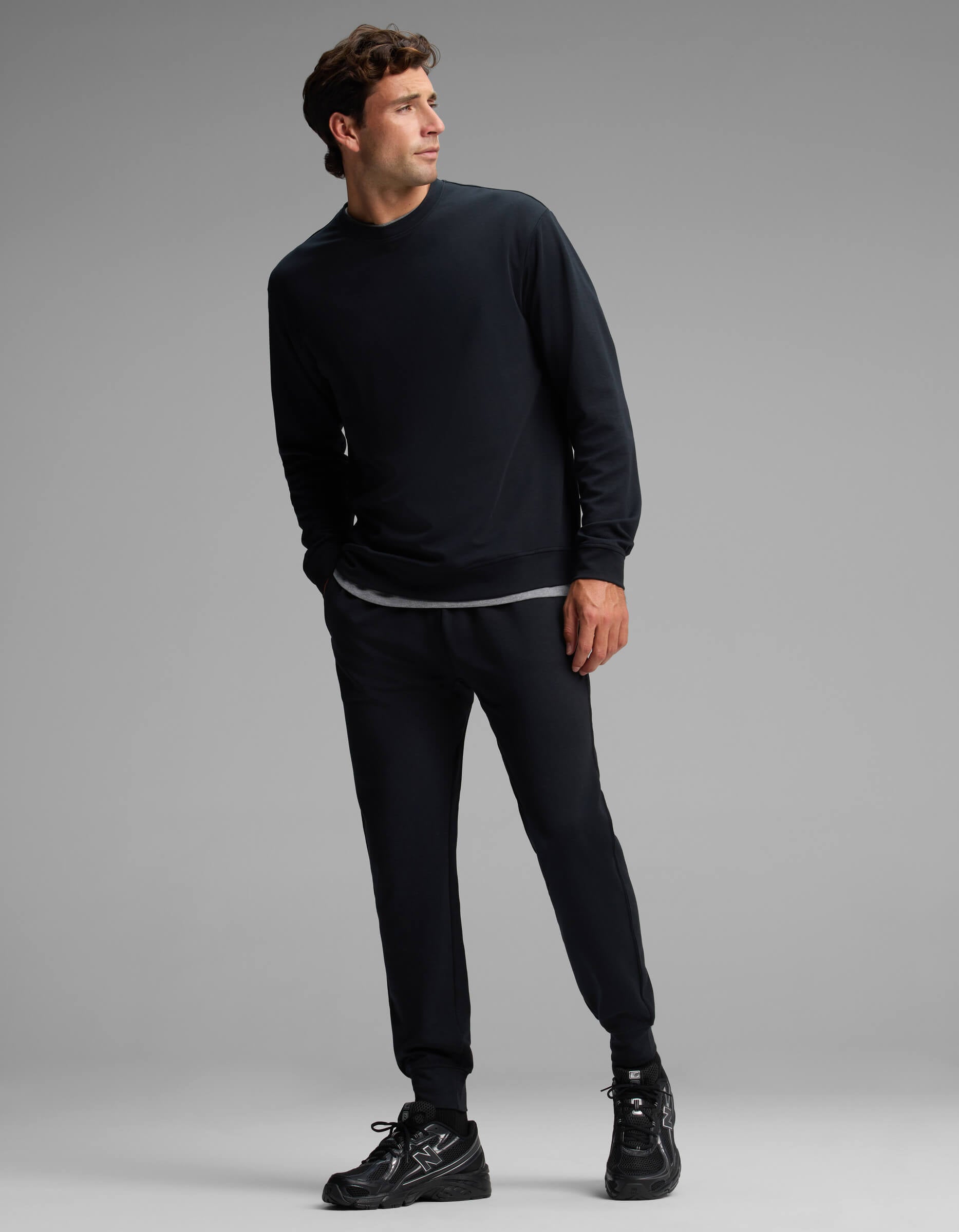 A man stands in a relaxed pose against a plain gray background, wearing the Cozy Earth Men's Versa Crewneck & Jogger Set in black, paired with black sneakers and a visible gray undershirt.|Color:Jet Black