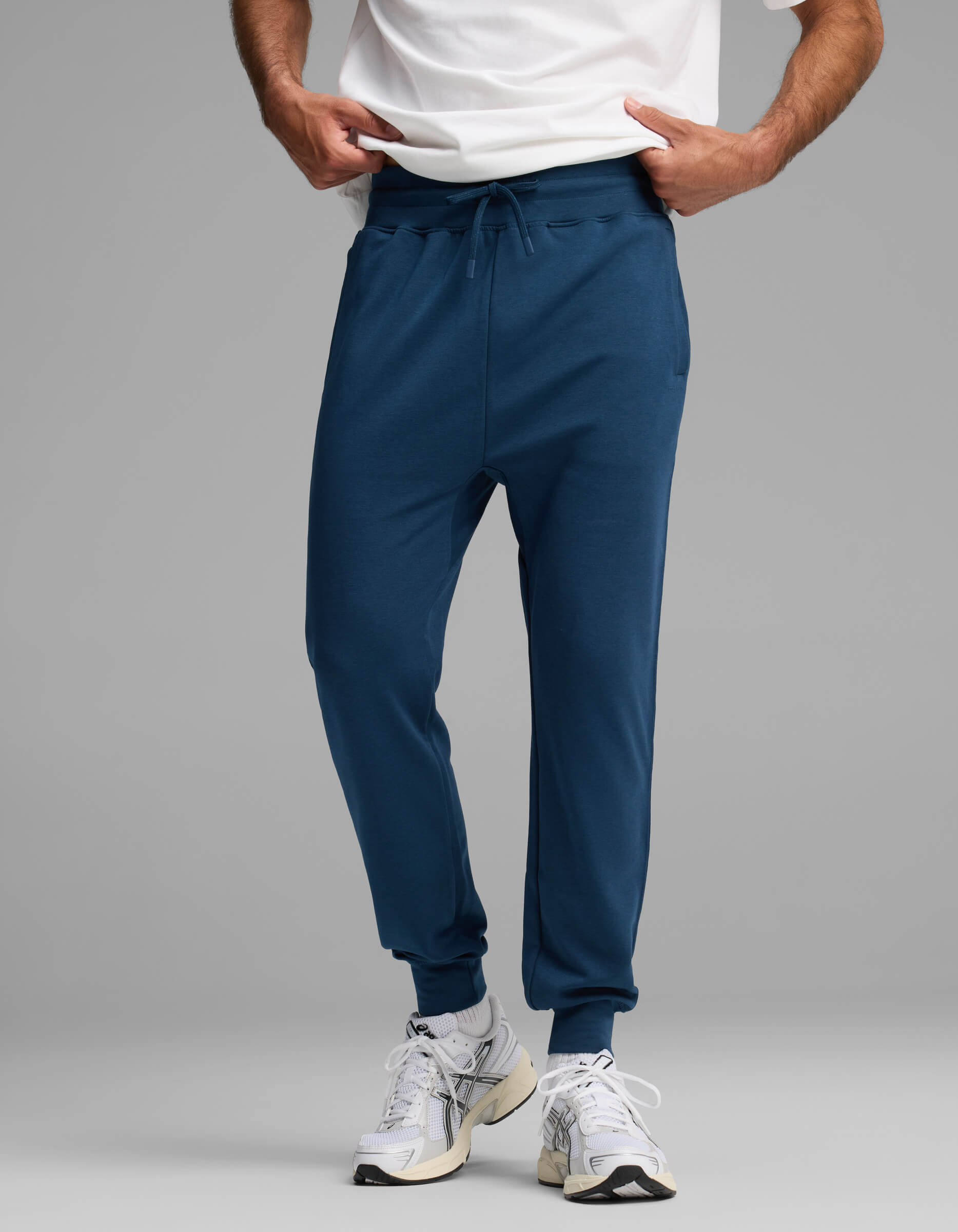 A person wearing a white shirt, Cozy Earth Men's Versa Jogger in blue, and white athletic shoes stands against a plain gray background with their hands in their pockets.|Color:Obsidian